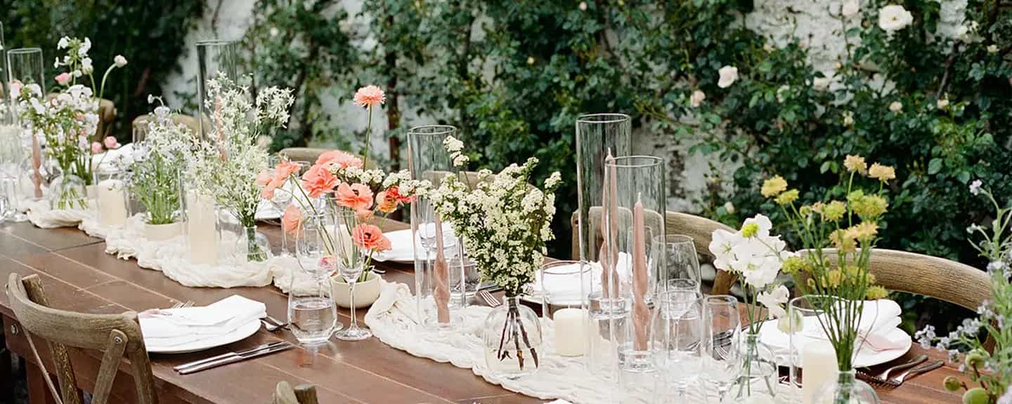 Long wooden table with bud vases of coral poppies, white spirea and sweet pea among taper candles, climbing roses behind
