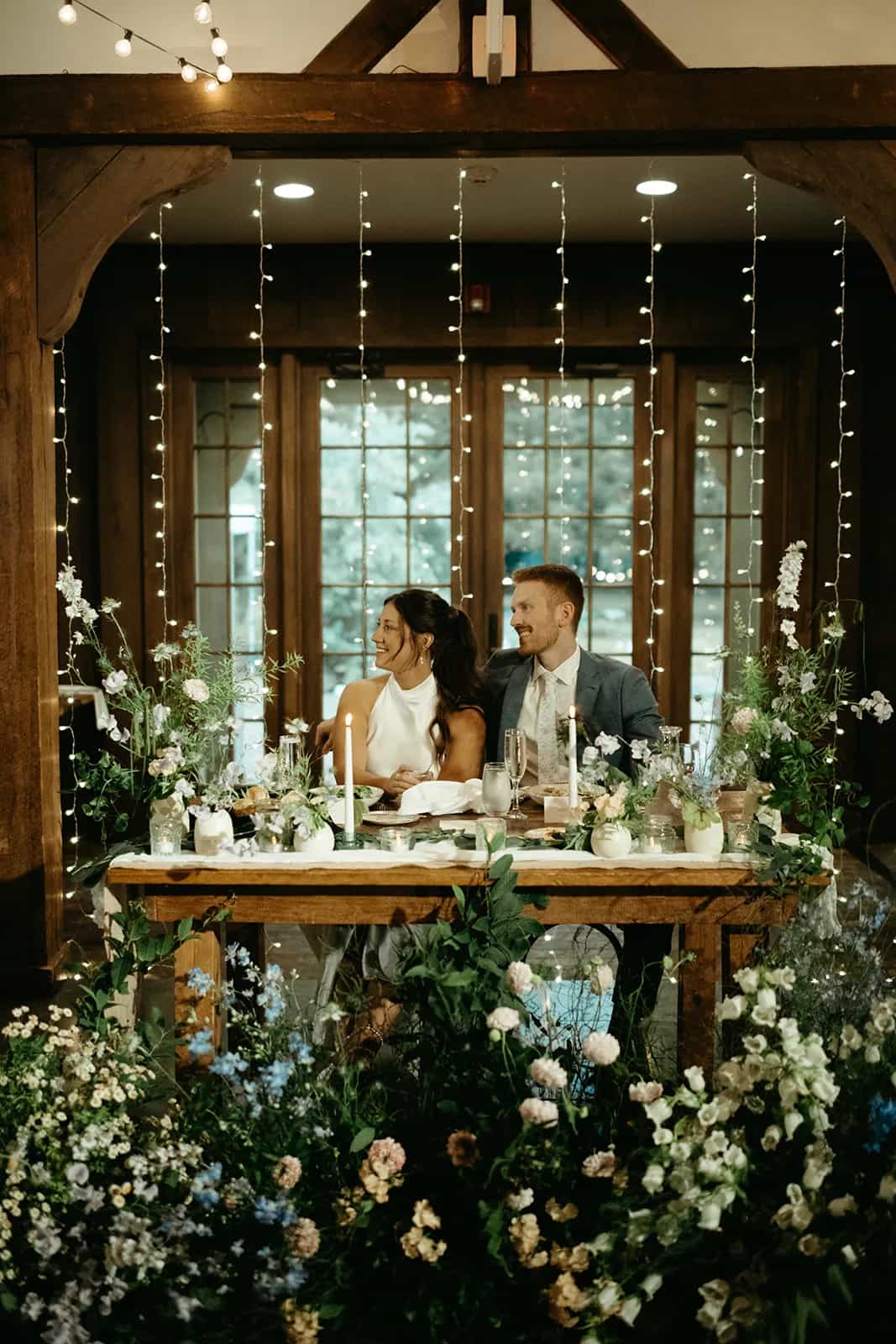 Shayna and Evan at the sweetheart table before a curtain of lights, bud vases and tapers on the runner, blooms massed below