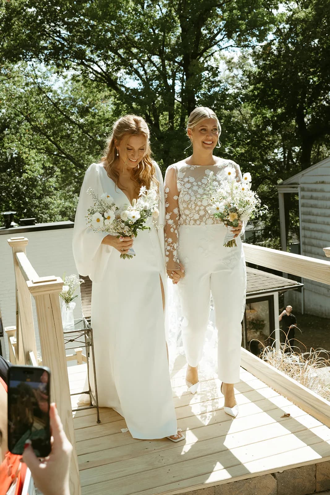 Chelsea and Jessica walk hand in hand across a sunlit deck, each holding a loose bouquet of white cosmos and daisies