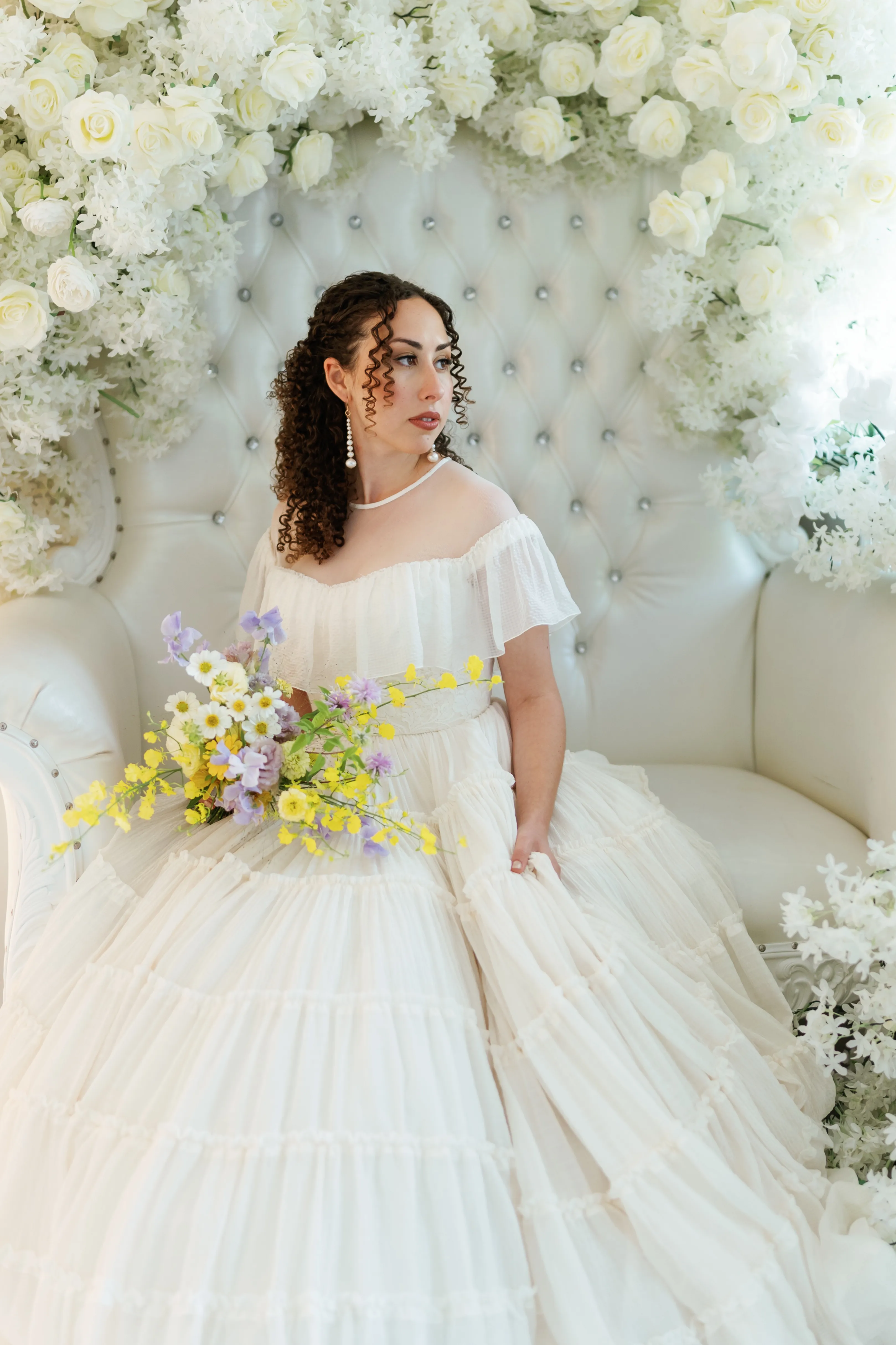 Seated bride beneath a canopy of white roses holds a bouquet of yellow oncidium orchids, lilac sweet pea and white cosmos