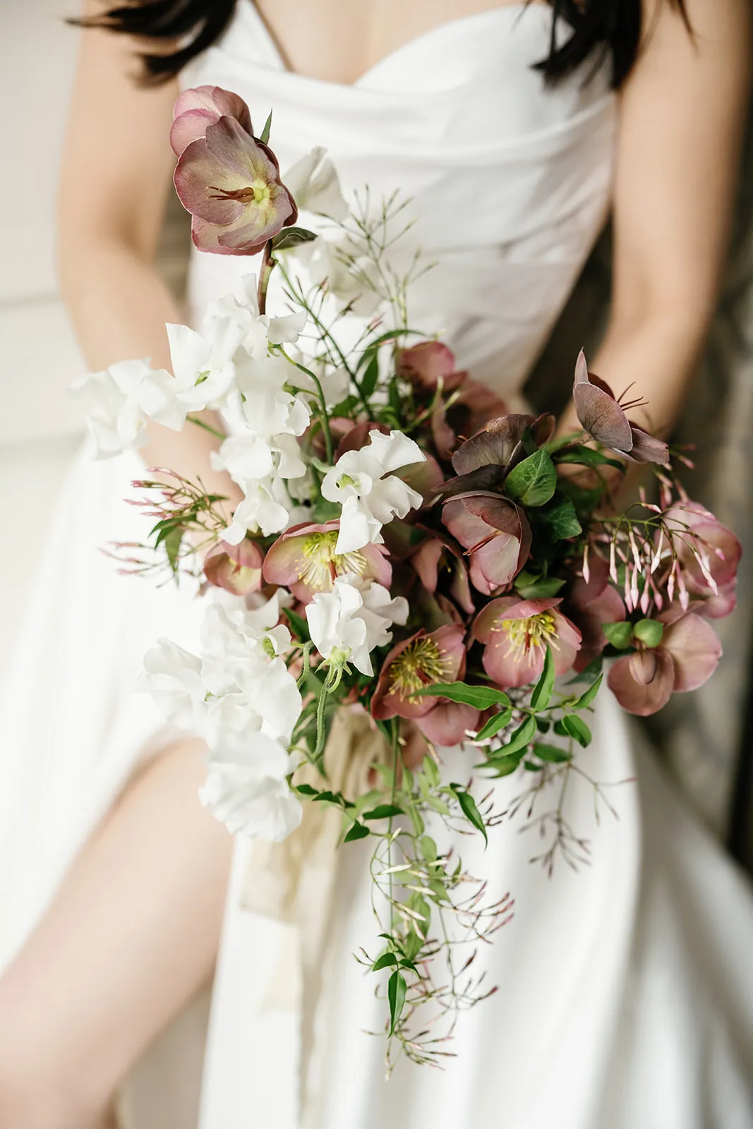Bride holds a cascading bouquet of dusky mauve hellebore, white sweet pea and trailing jasmine vine tied with silk ribbon