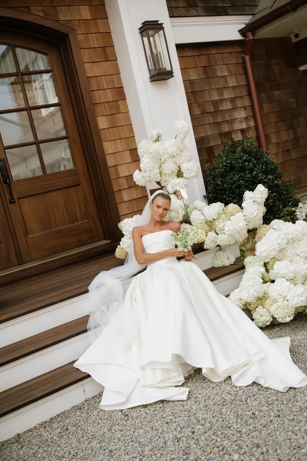 Lindsey sits on the steps of a shingled porch among sculptural mounds of white hydrangea, holding a small tweedia bouquet
