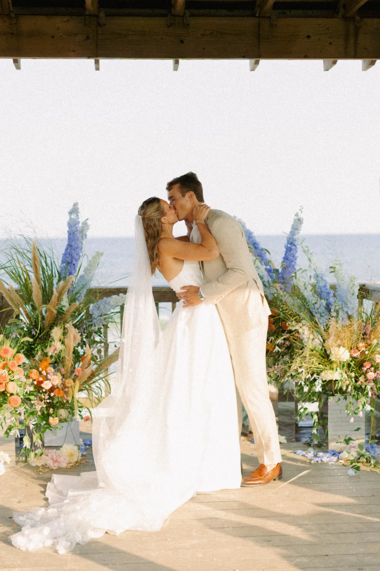 Talea and Erich kiss beneath a wooden pavilion by the sea, framed by tall blue delphinium, orange dahlias and pale grasses