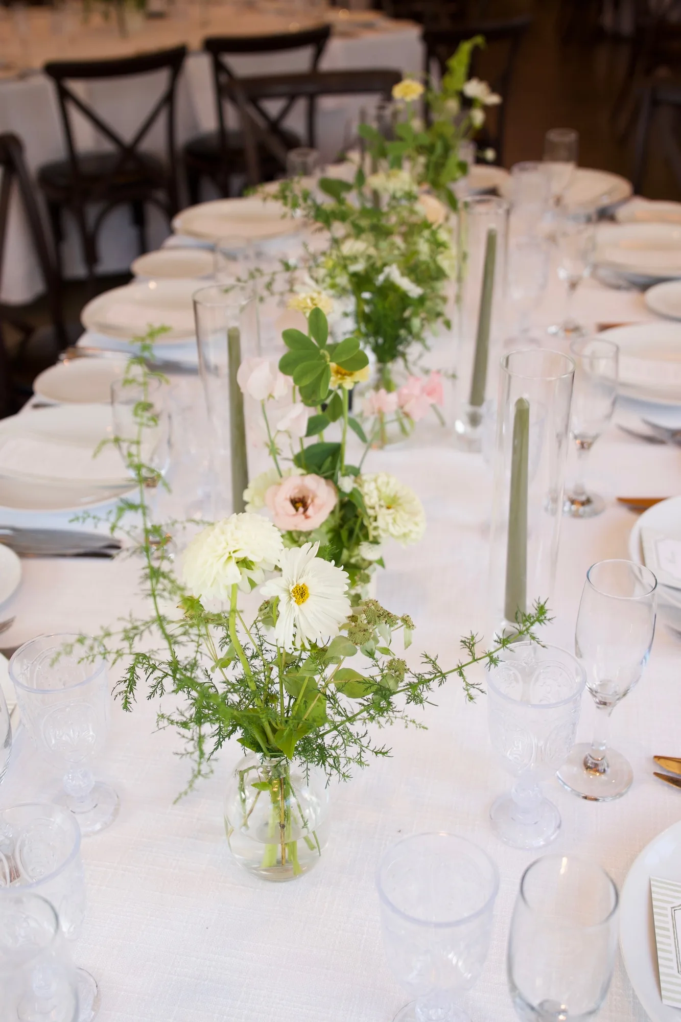 Bud vases of white dahlias, blush lisianthus and sweet pea run down a long white table between sage taper candles