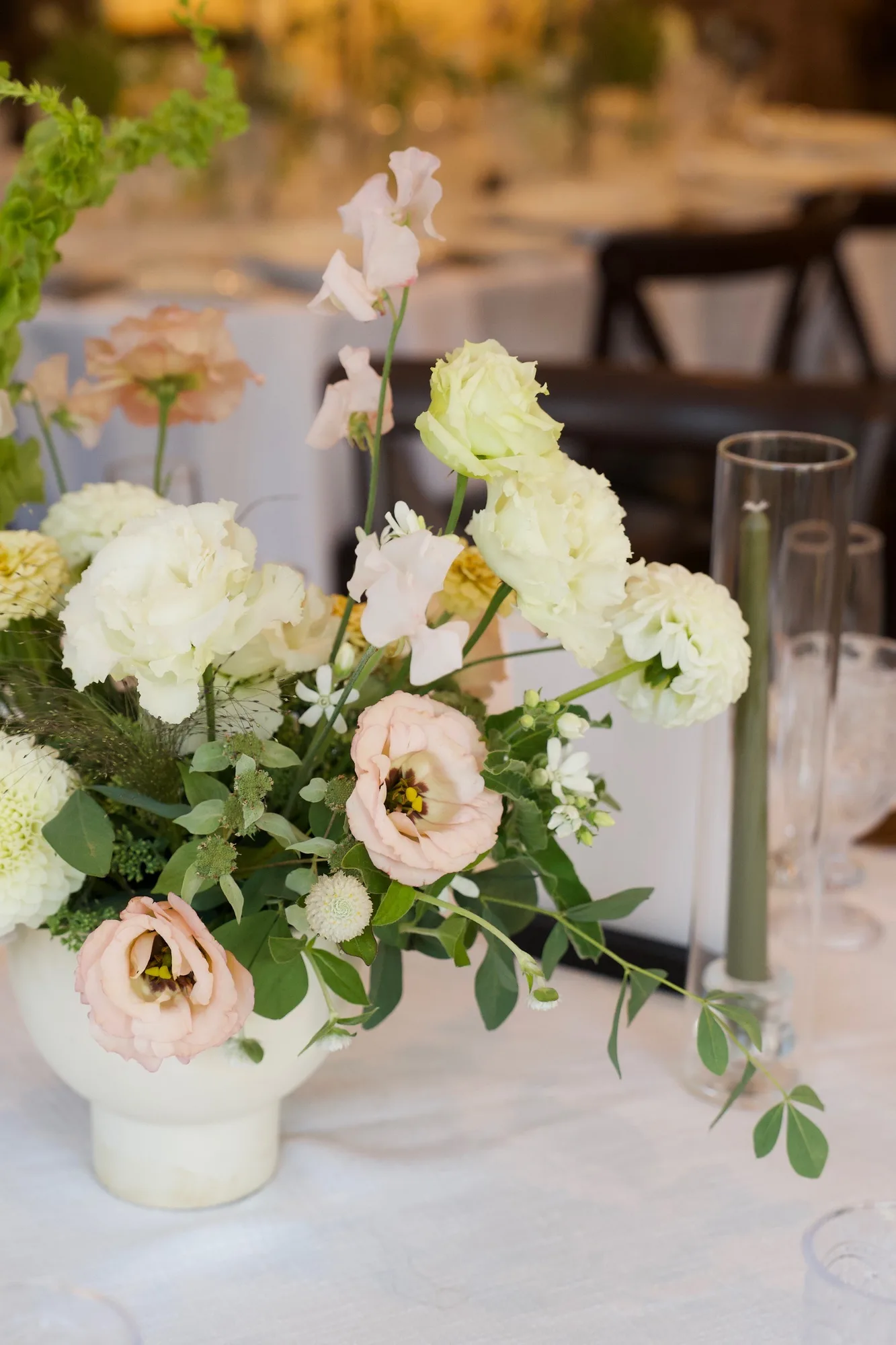 Footed white vessel of blush lisianthus, cream ranunculus, sweet pea and dahlias beside a sage taper on white linen