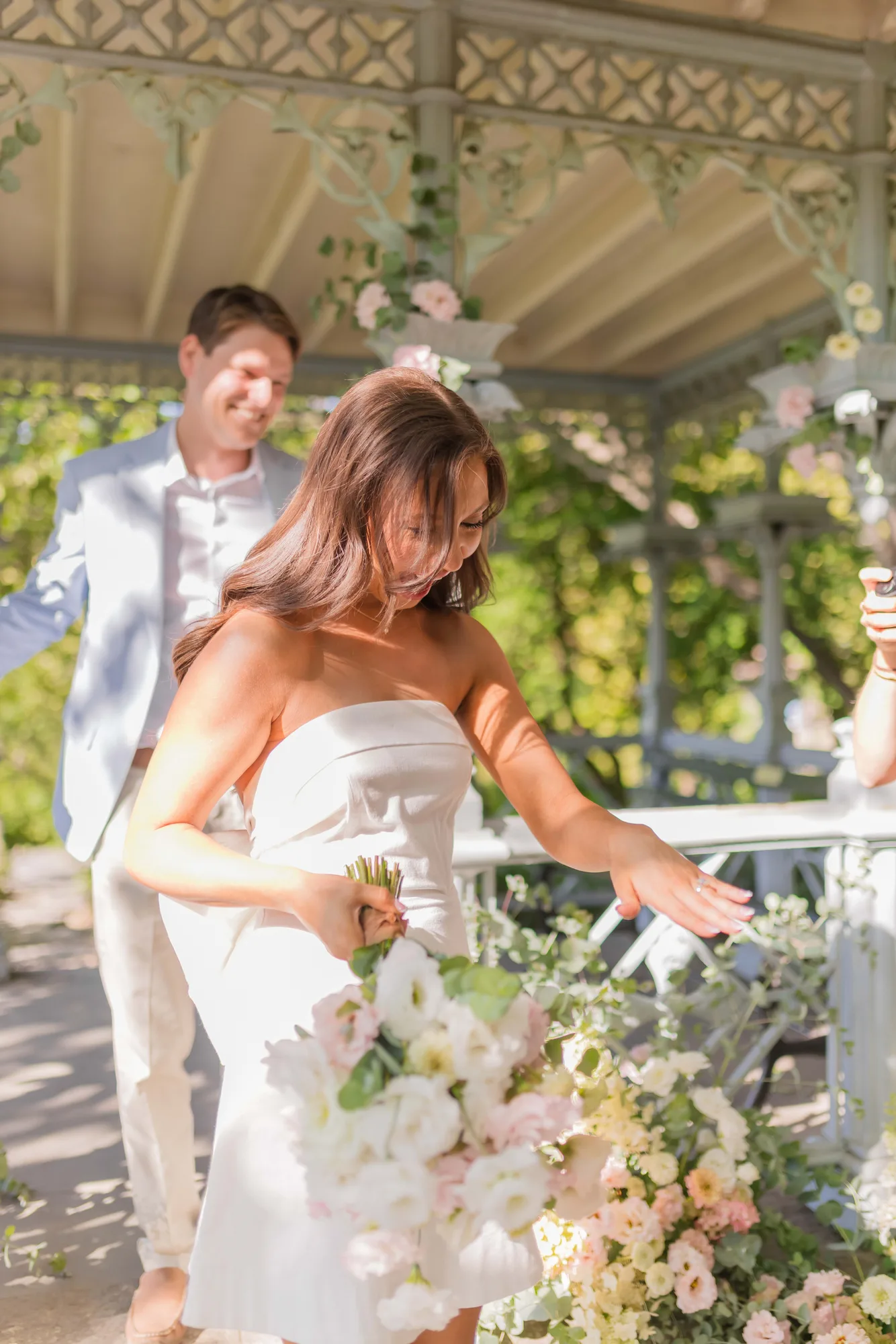 Lauren looks down at the ring on her hand, bouquet in the other, Will smiling behind her beside a blush ground arrangement