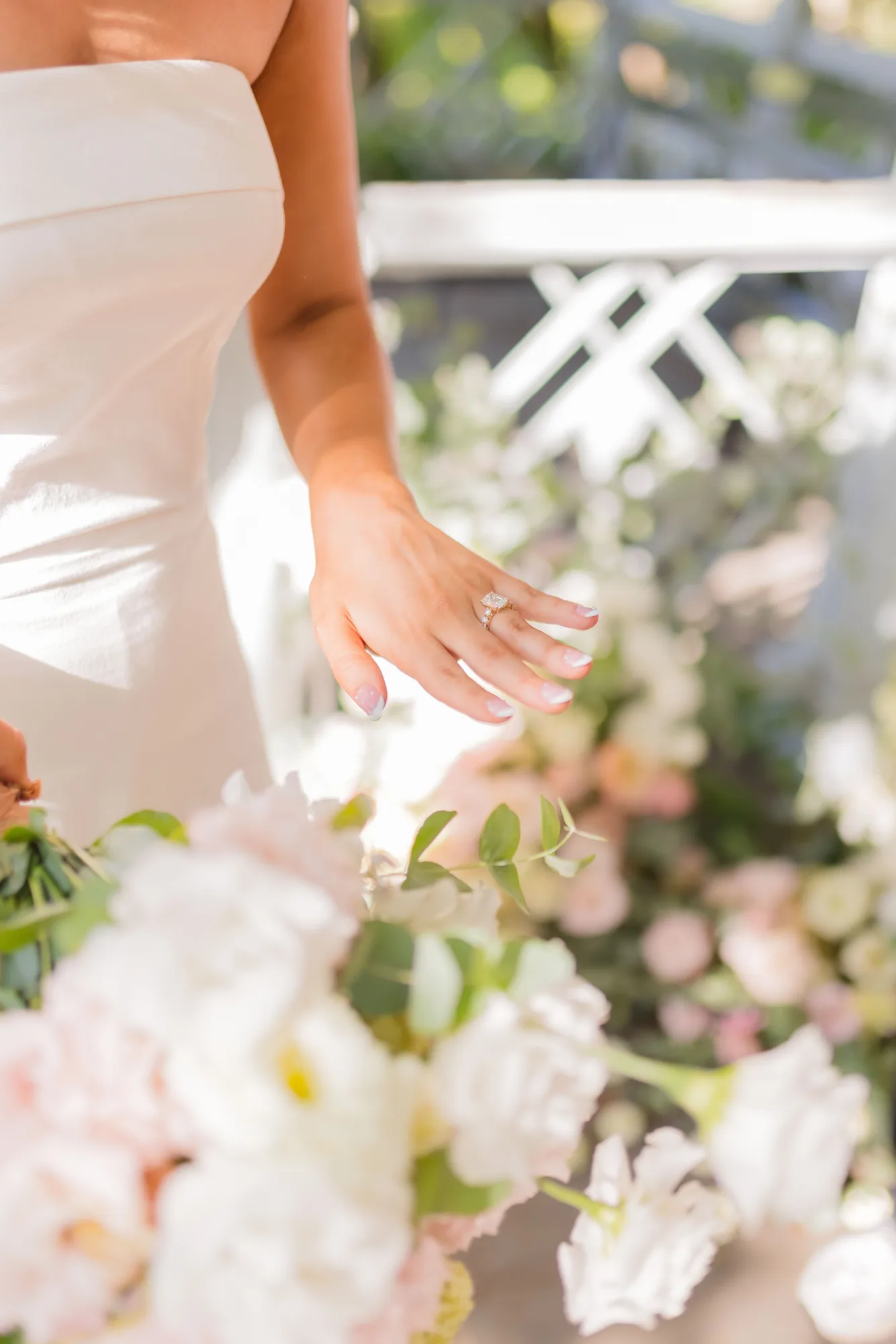 Lauren's hand with an emerald-cut ring hovers above a soft blur of blush and cream blooms in her bouquet