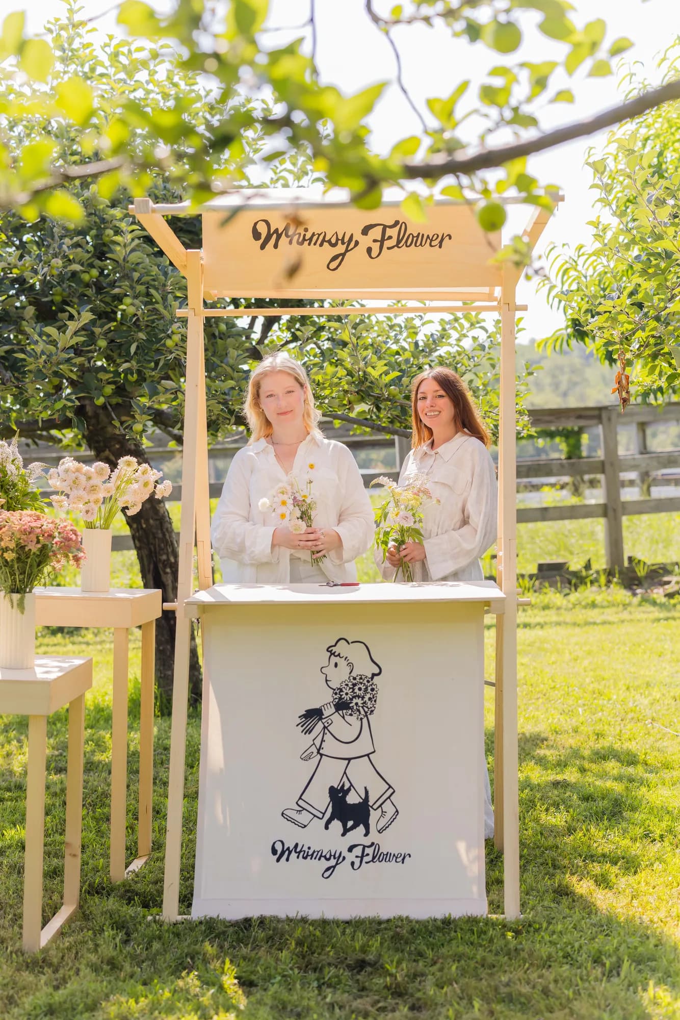 Two women behind a canvas-fronted wooden flower bar under an apple tree, each holding a small hand-tied bouquet