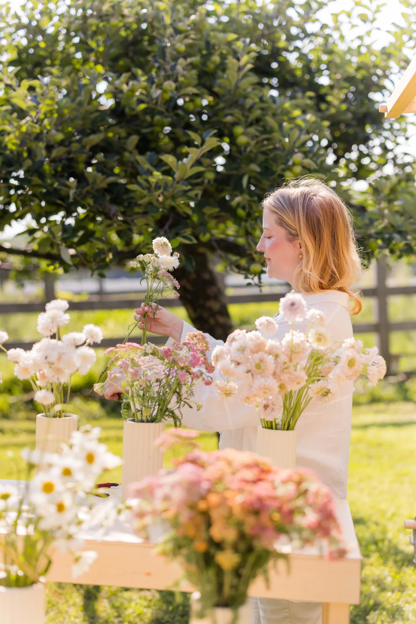 A guest in white linen lifts white scabiosa and pink yarrow from fluted vases on a plywood flower bar beneath an apple tree