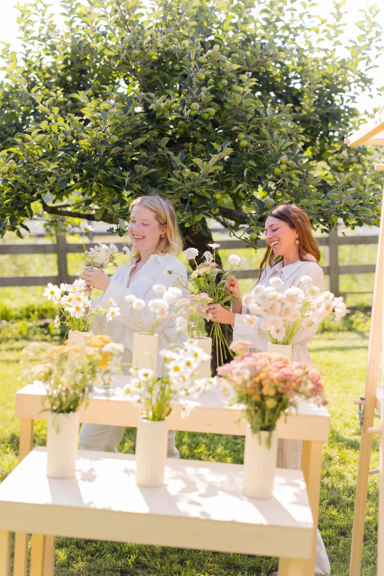 Two women laugh behind a plywood flower bar, gathering daisies, scabiosa and pink yarrow from white fluted vases