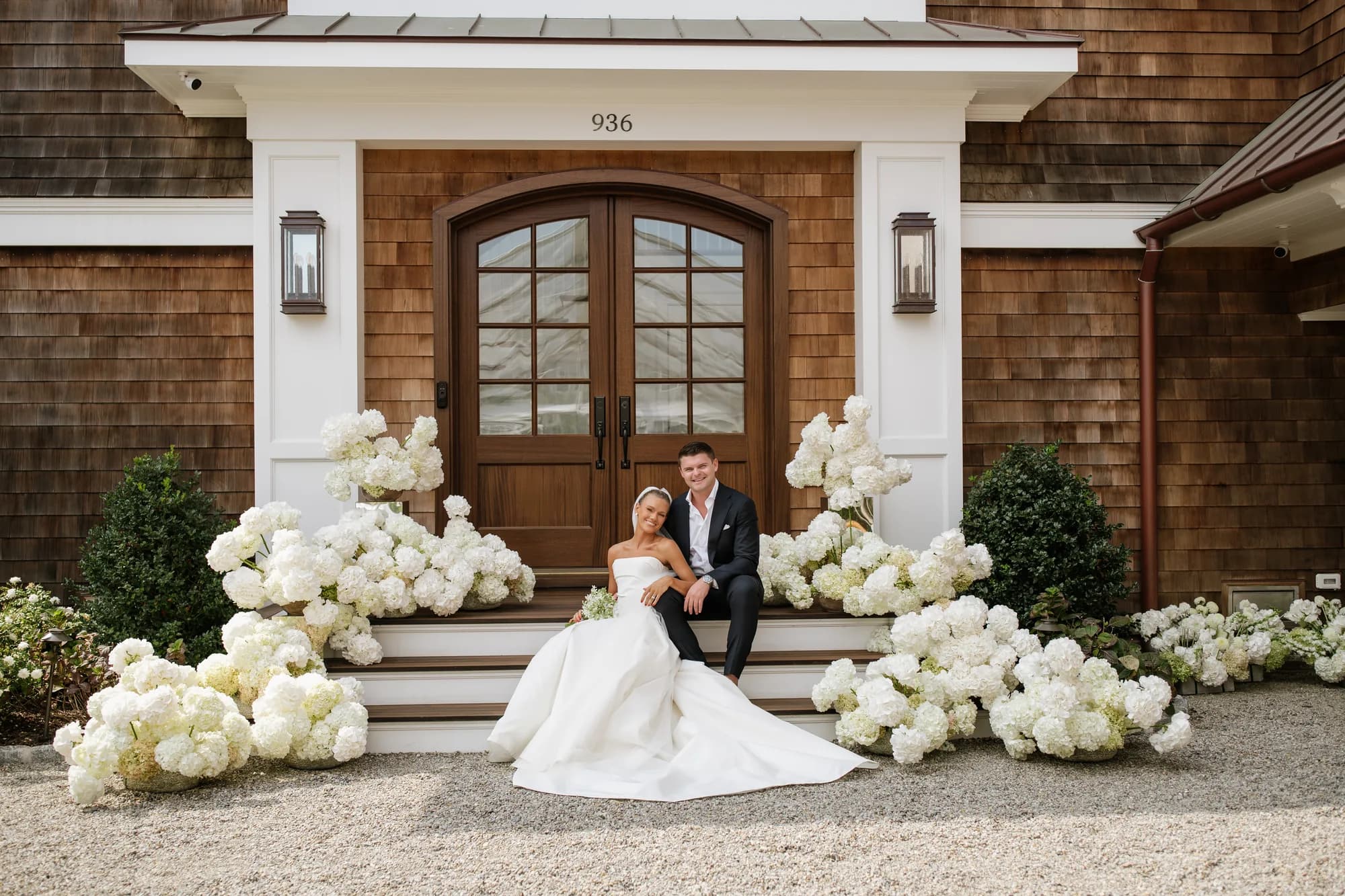 Wide view of Lindsey and Finn seated on the steps, white hydrangea pooled across the stairs and onto the gravel