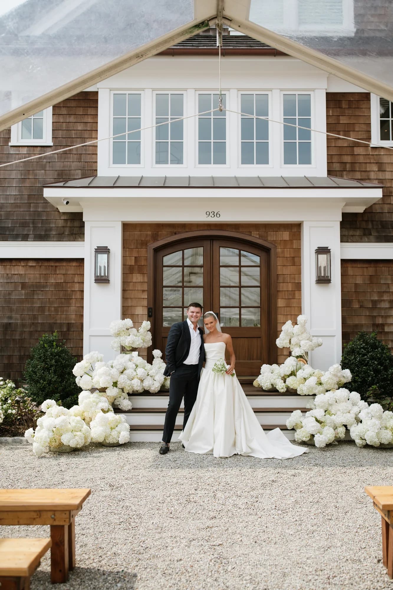 Lindsey and Finn stand before the front steps under a clear tent, white hydrangea banked on both sides