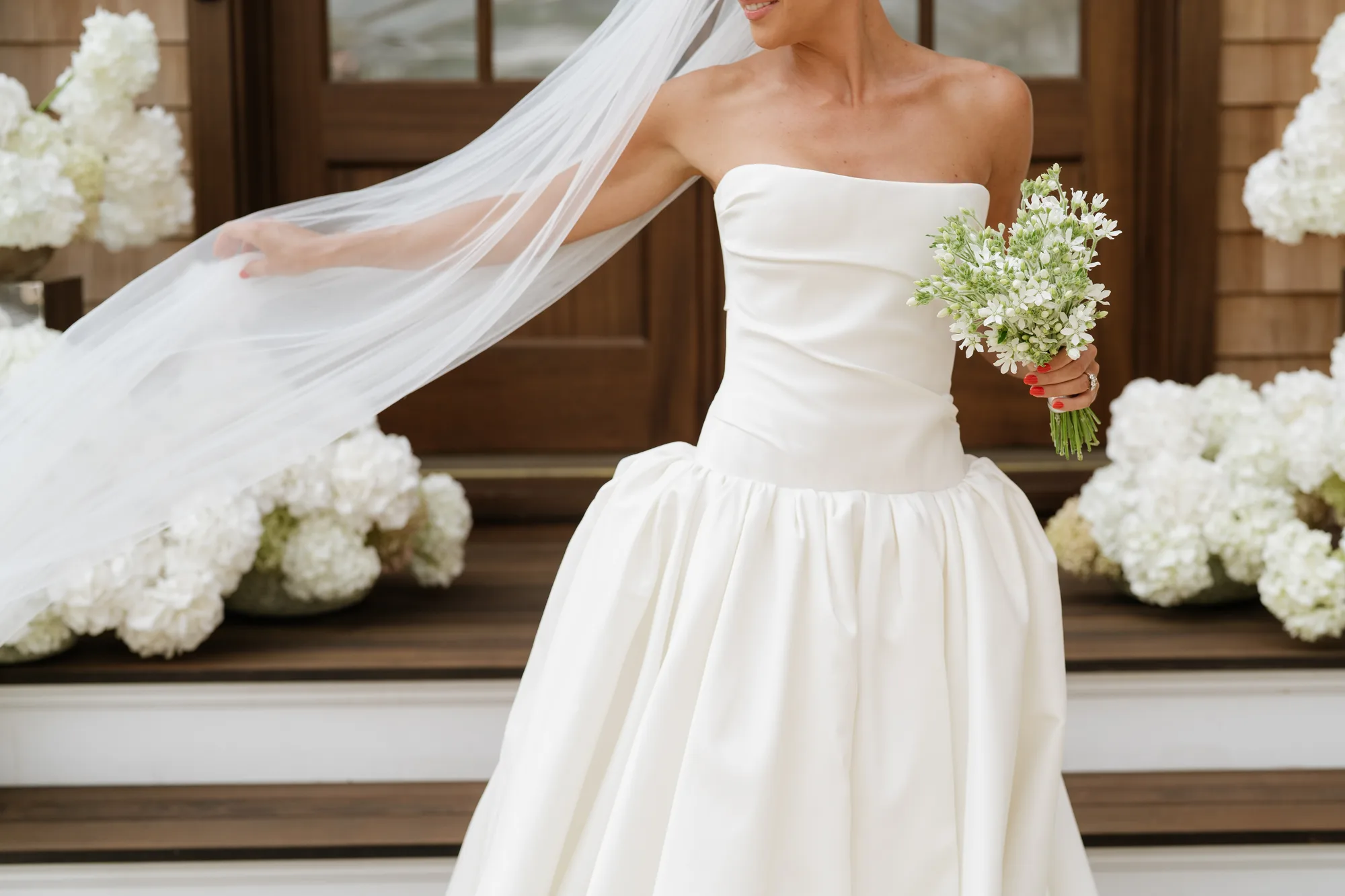 Lindsey lifts her veil in one hand and her tweedia bouquet in the other, hydrangea on the steps behind her