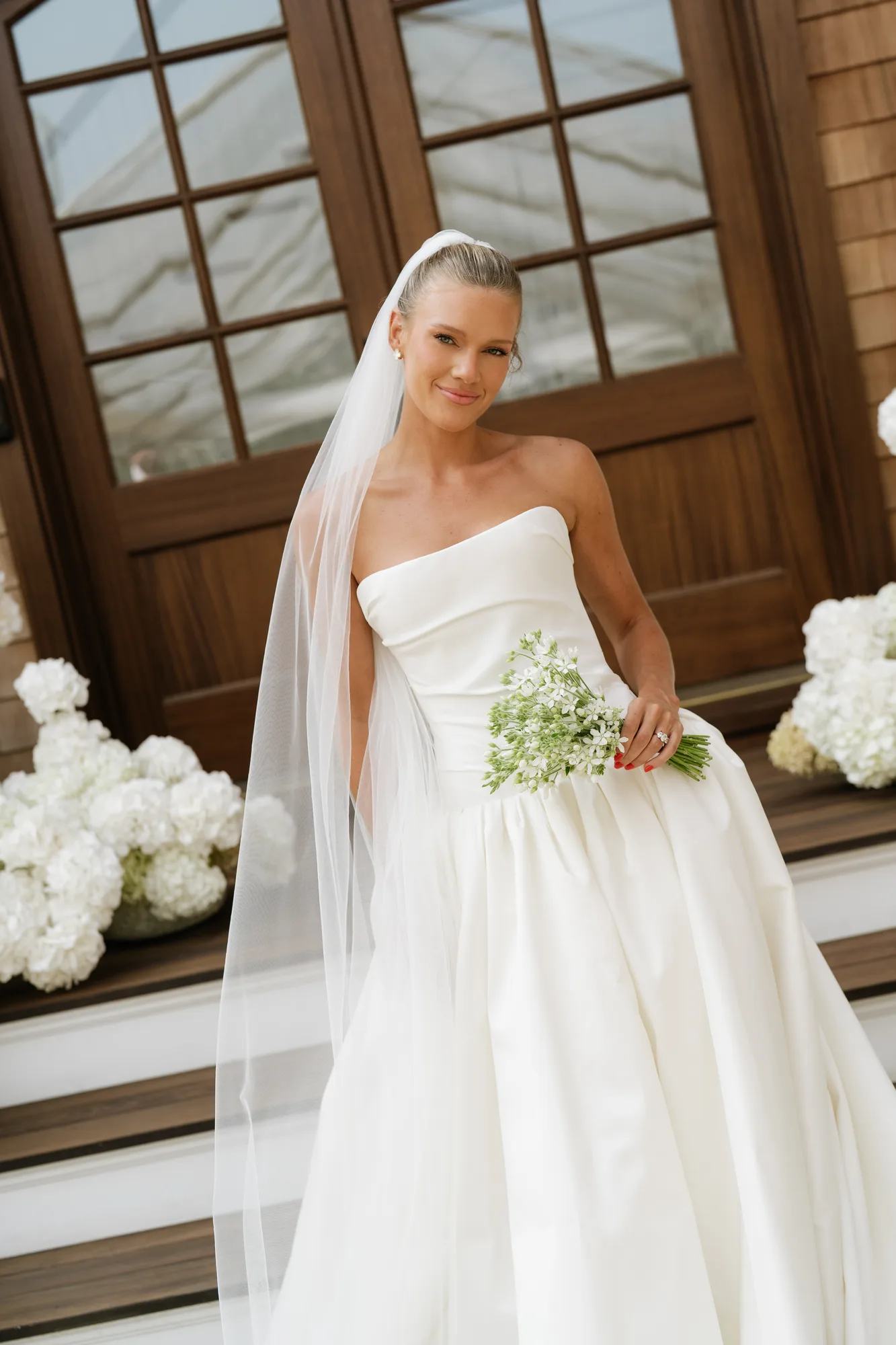 Lindsey in her veil on the front steps holding her tweedia bouquet, white hydrangea banked at either side
