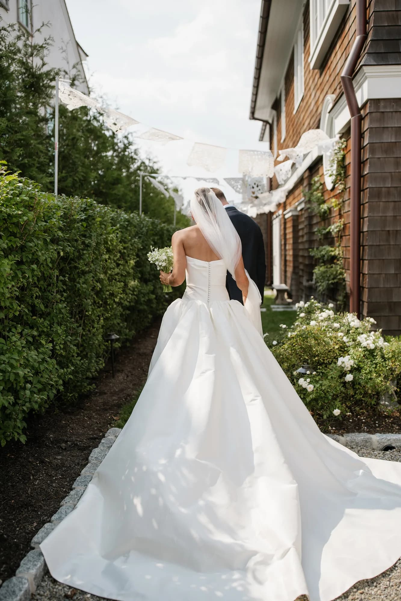 Lindsey and Finn walk a hedge-lined path beneath white paper banners, her tweedia bouquet in hand
