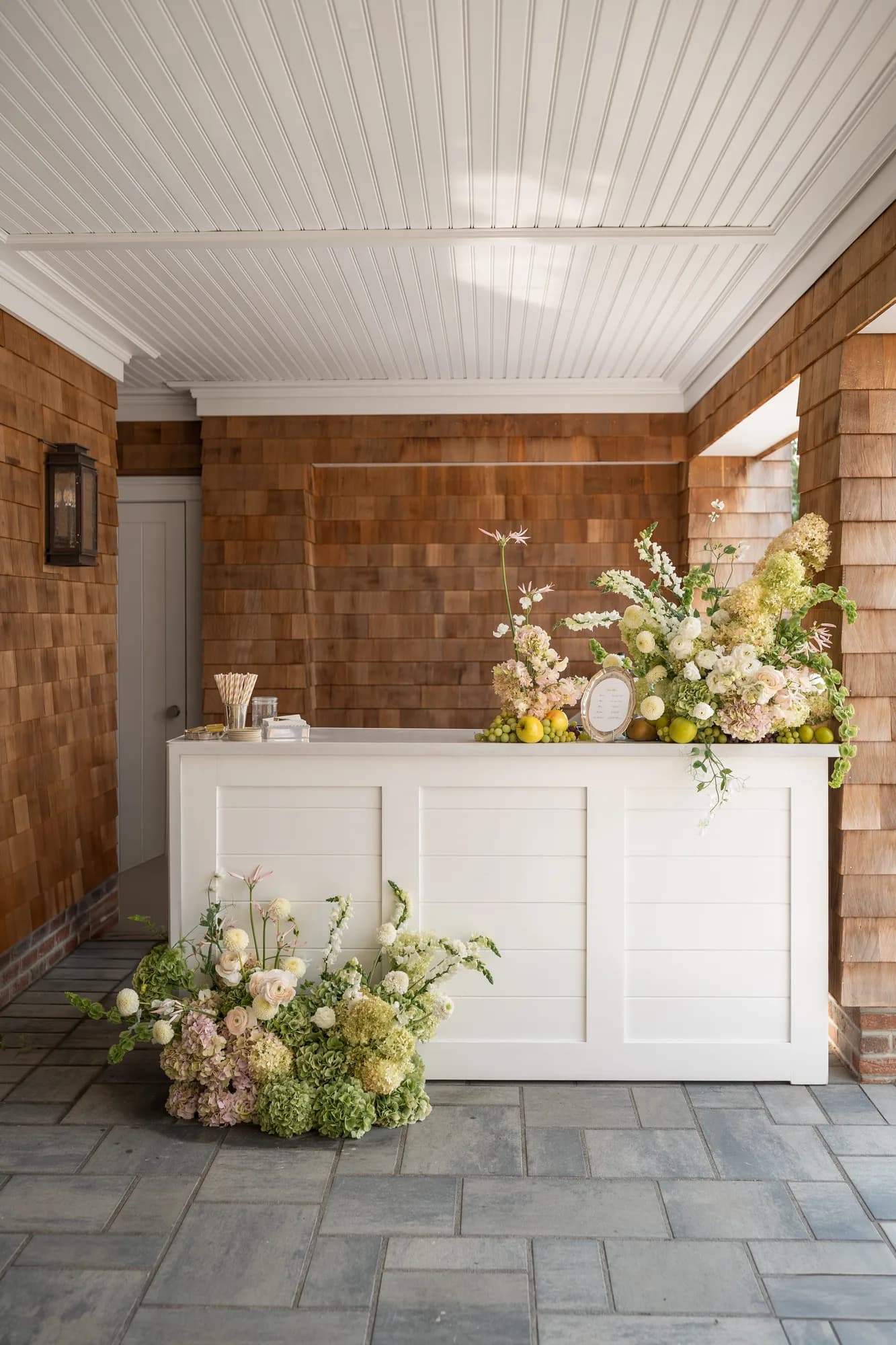 A white paneled bar in a shingled breezeway, a cascading arrangement on top and a low one at its base