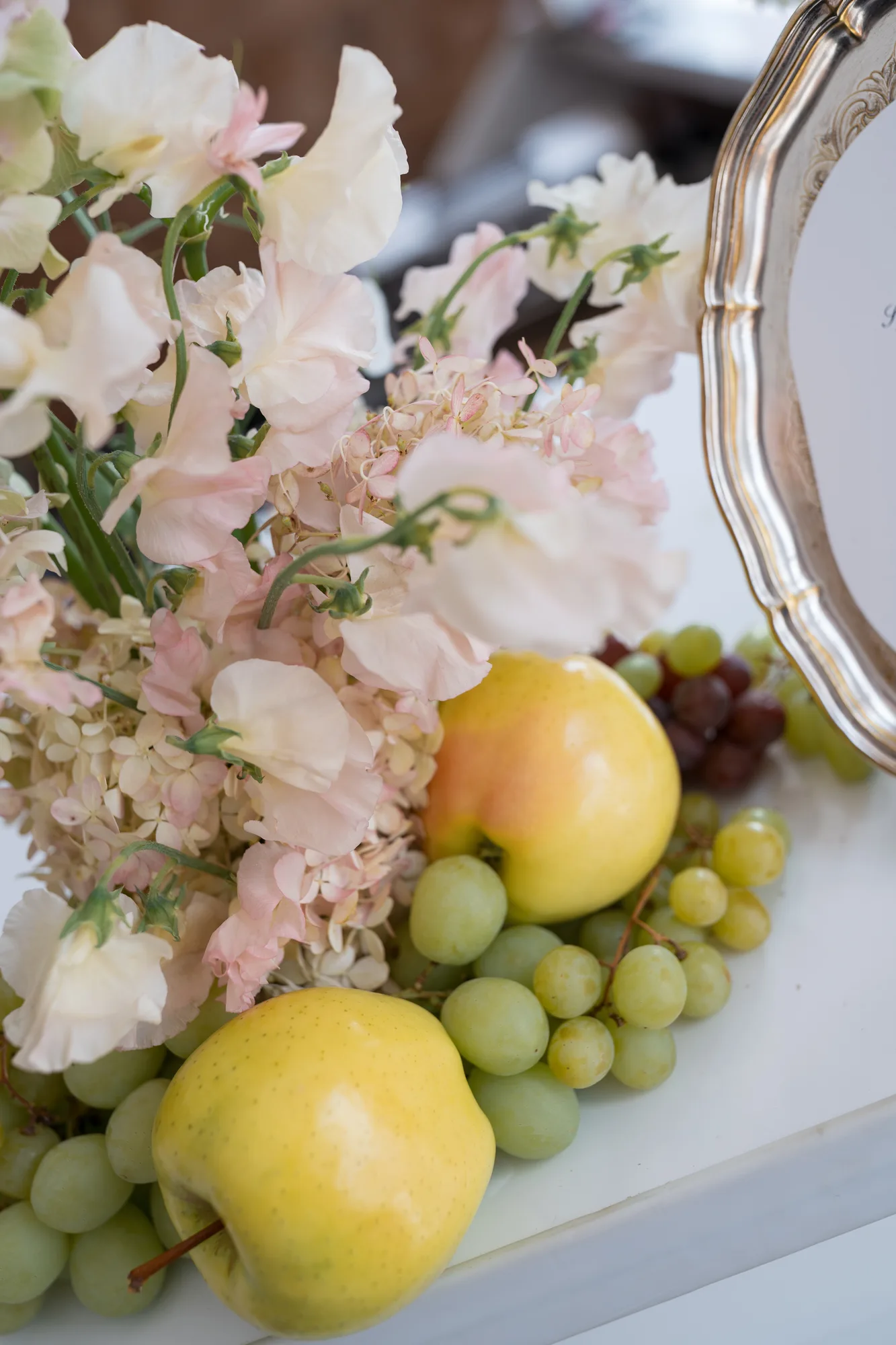 Blush sweet pea and faded hydrangea lean over yellow apples and green grapes at the edge of a silver tray