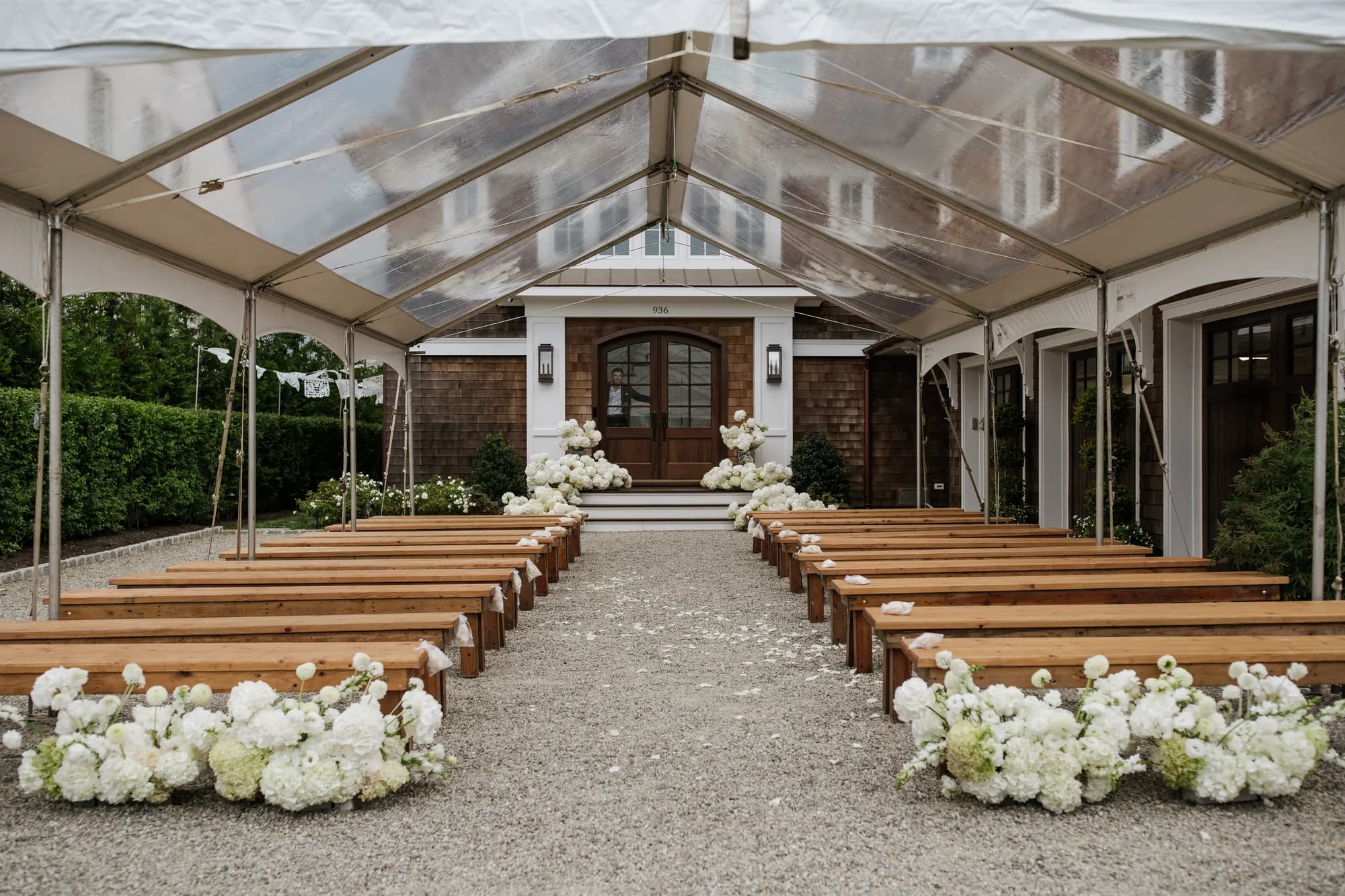 Wooden benches line a gravel aisle under a clear tent, white arrangements at each end and hydrangea on the steps