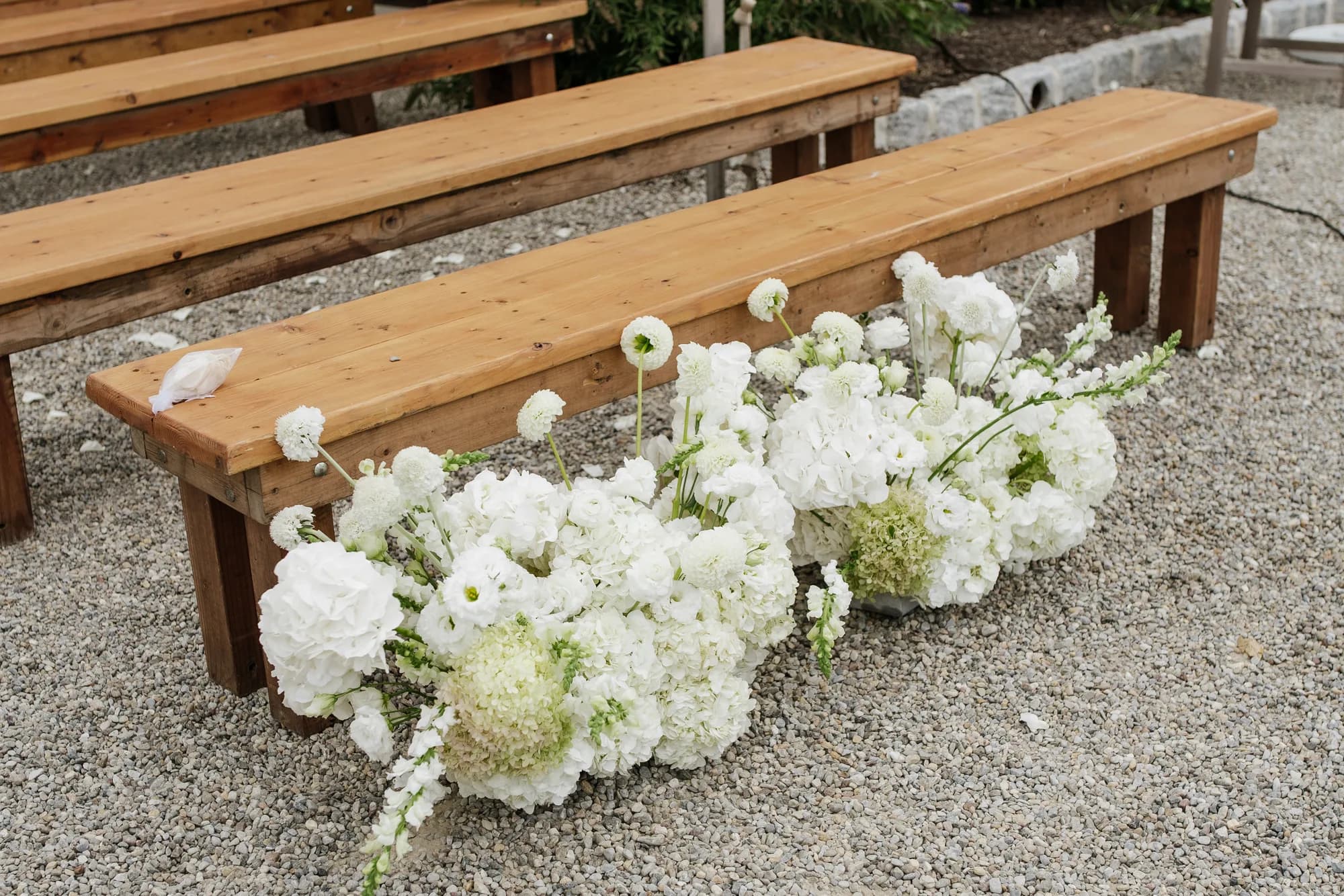 A white arrangement of hydrangea, dahlias, lisianthus and snapdragon set at the end of a wooden bench on gravel
