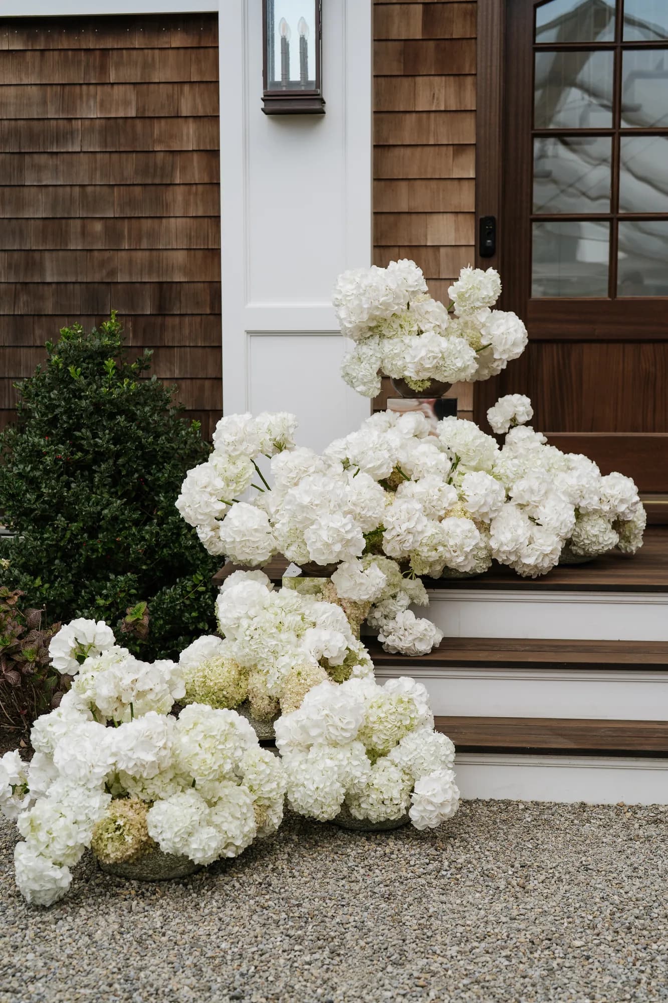 White hydrangea spills down the front steps beside boxwood, one urn raised on a mirrored plinth