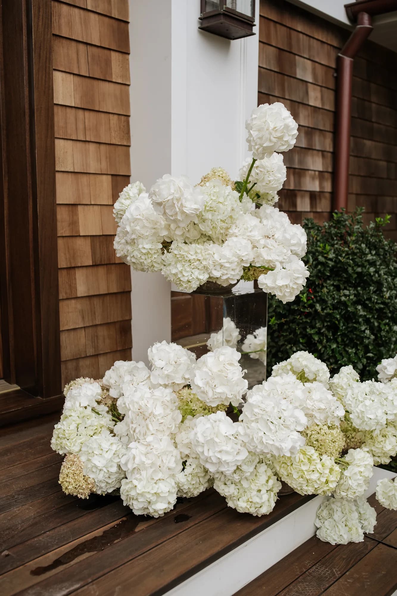 A footed bowl of white hydrangea on a mirrored cube, more hydrangea heads pooled across the decking below