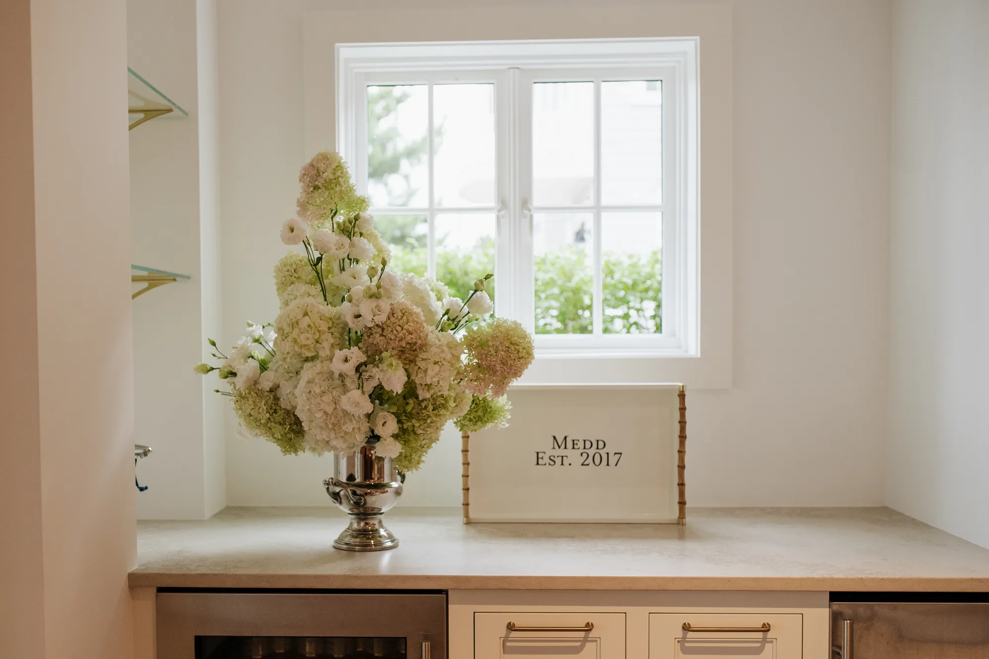 A silver trophy urn of limelight hydrangea and white lisianthus on a stone counter before a bright window