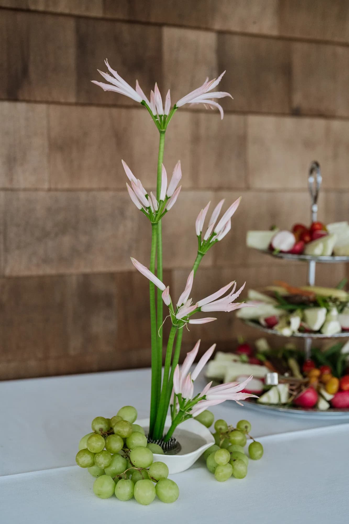 Pale pink nerine stems rise from a low white dish ringed with green grapes, an ikebana-style arrangement