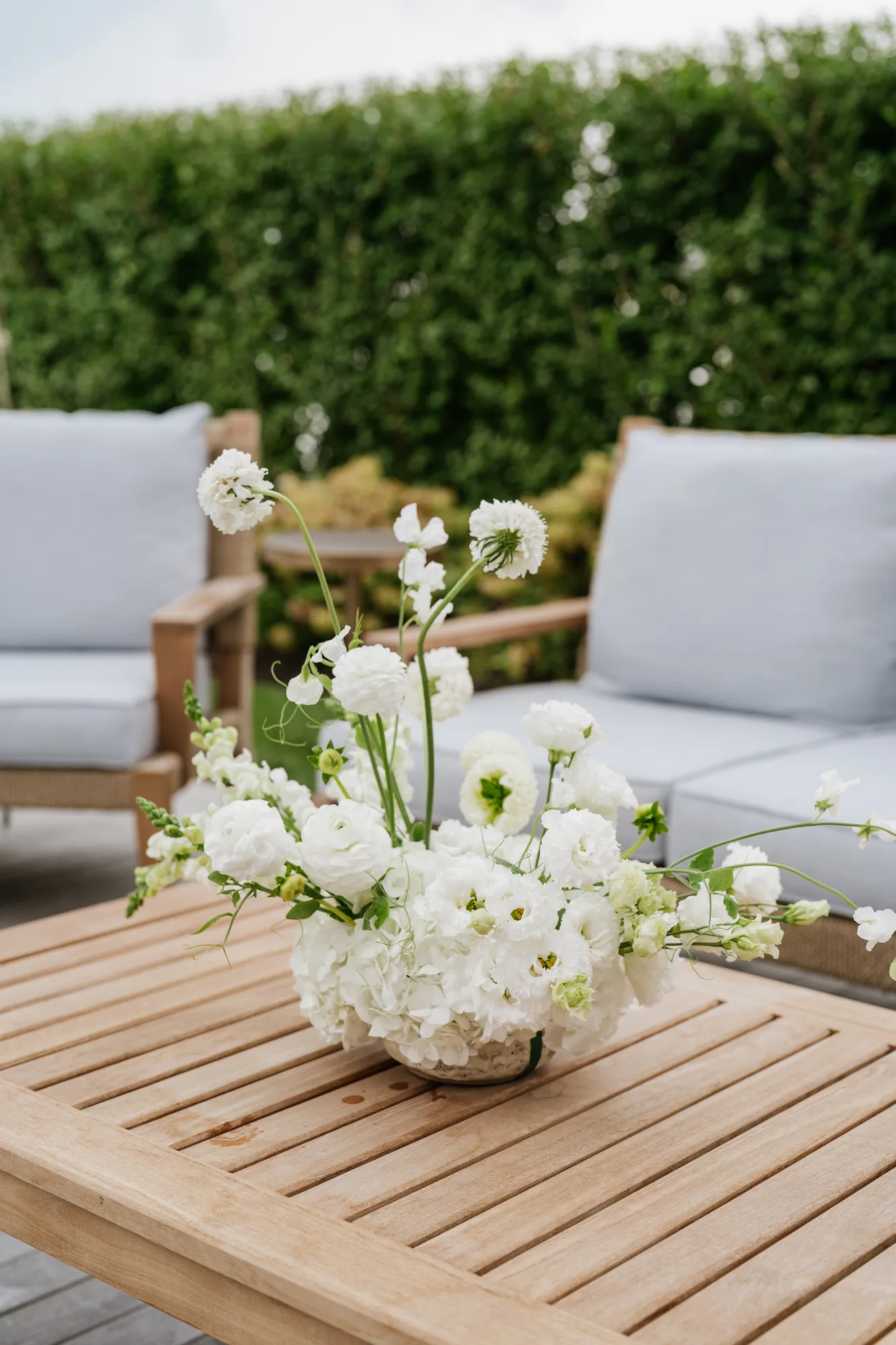 A low table arrangement of white hydrangea, ranunculus, lisianthus and scabiosa on a teak table by outdoor seating