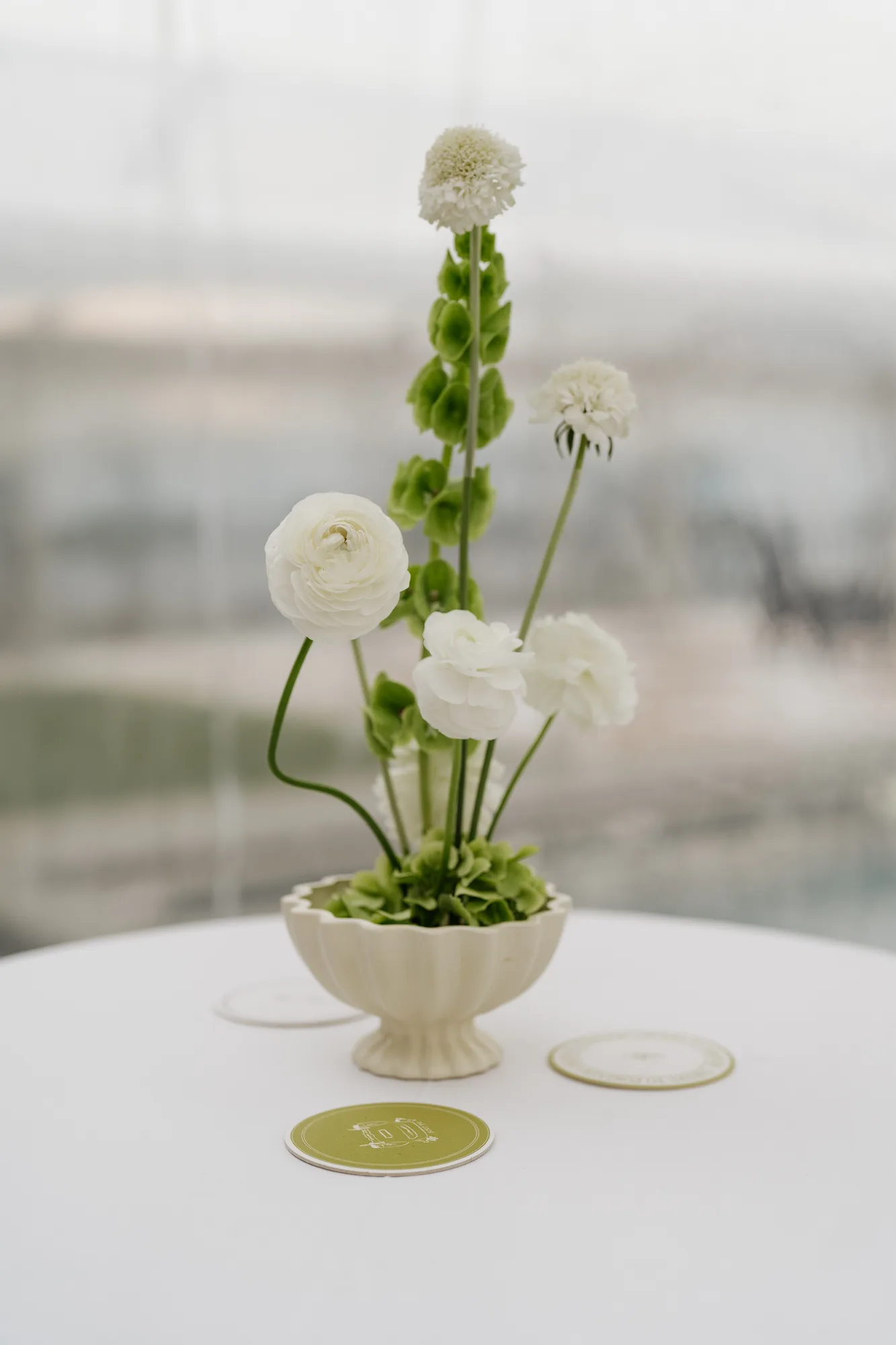 An ikebana-style table arrangement of white ranunculus, bells of Ireland and scabiosa in a cream fluted compote