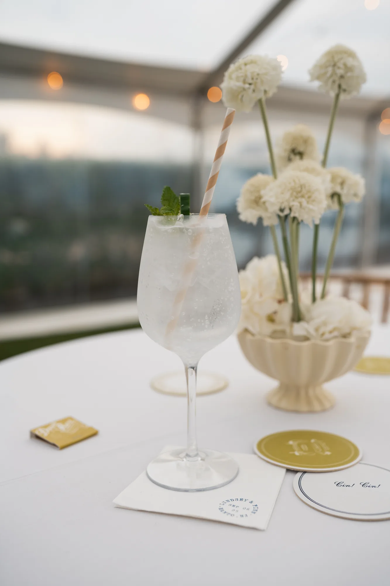 A spritz with a striped straw beside white scabiosa rising from a cream compote on a cocktail table