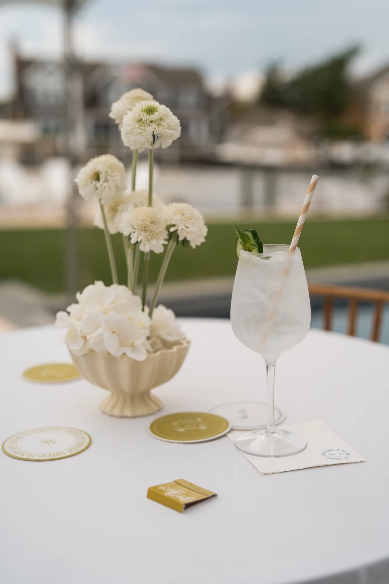 A cocktail table set with white scabiosa in a cream fluted compote, a spritz, chartreuse coasters and a matchbook