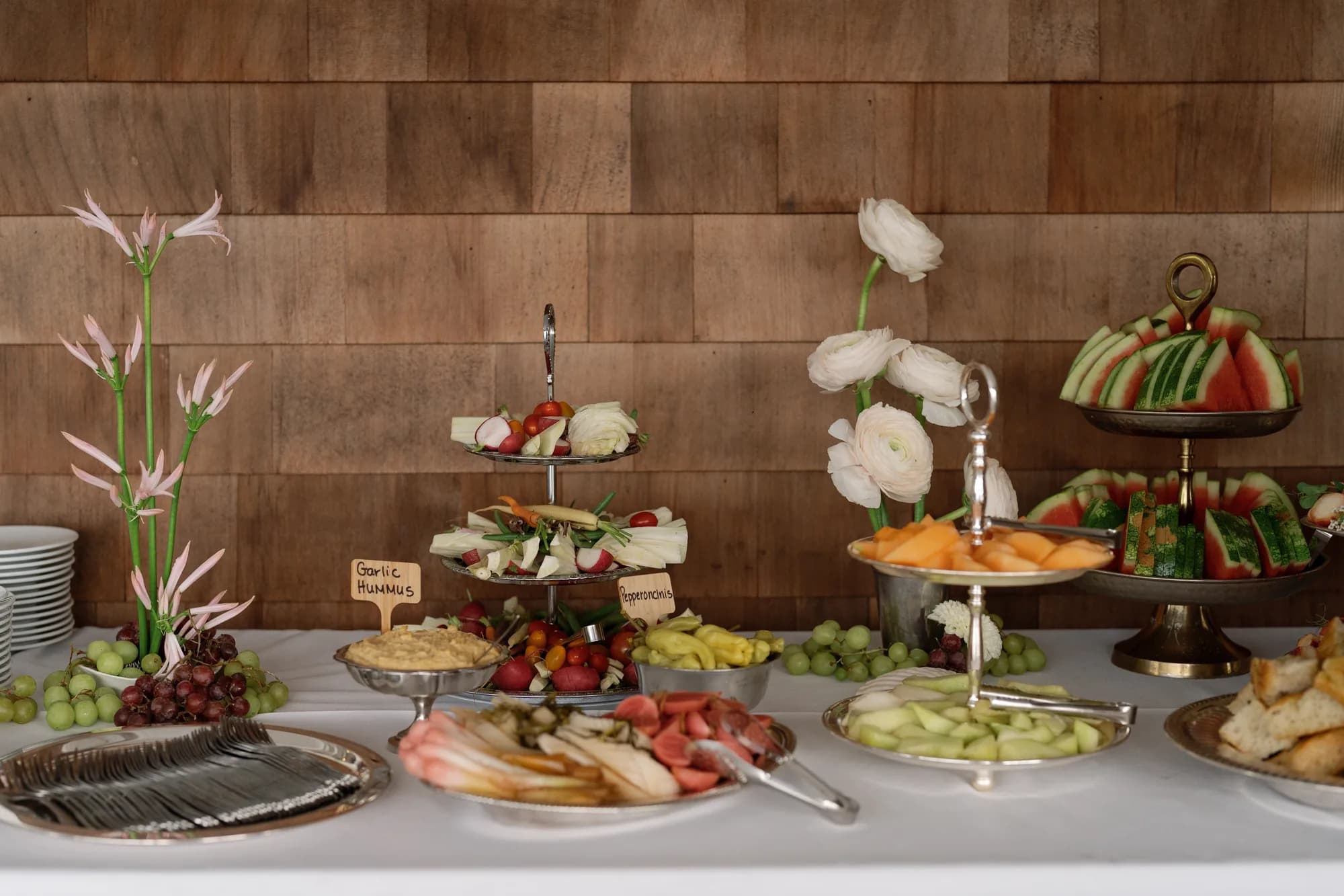 A buffet against a shingled wall with tiered trays of crudite and melon, pale nerine and white ranunculus stems