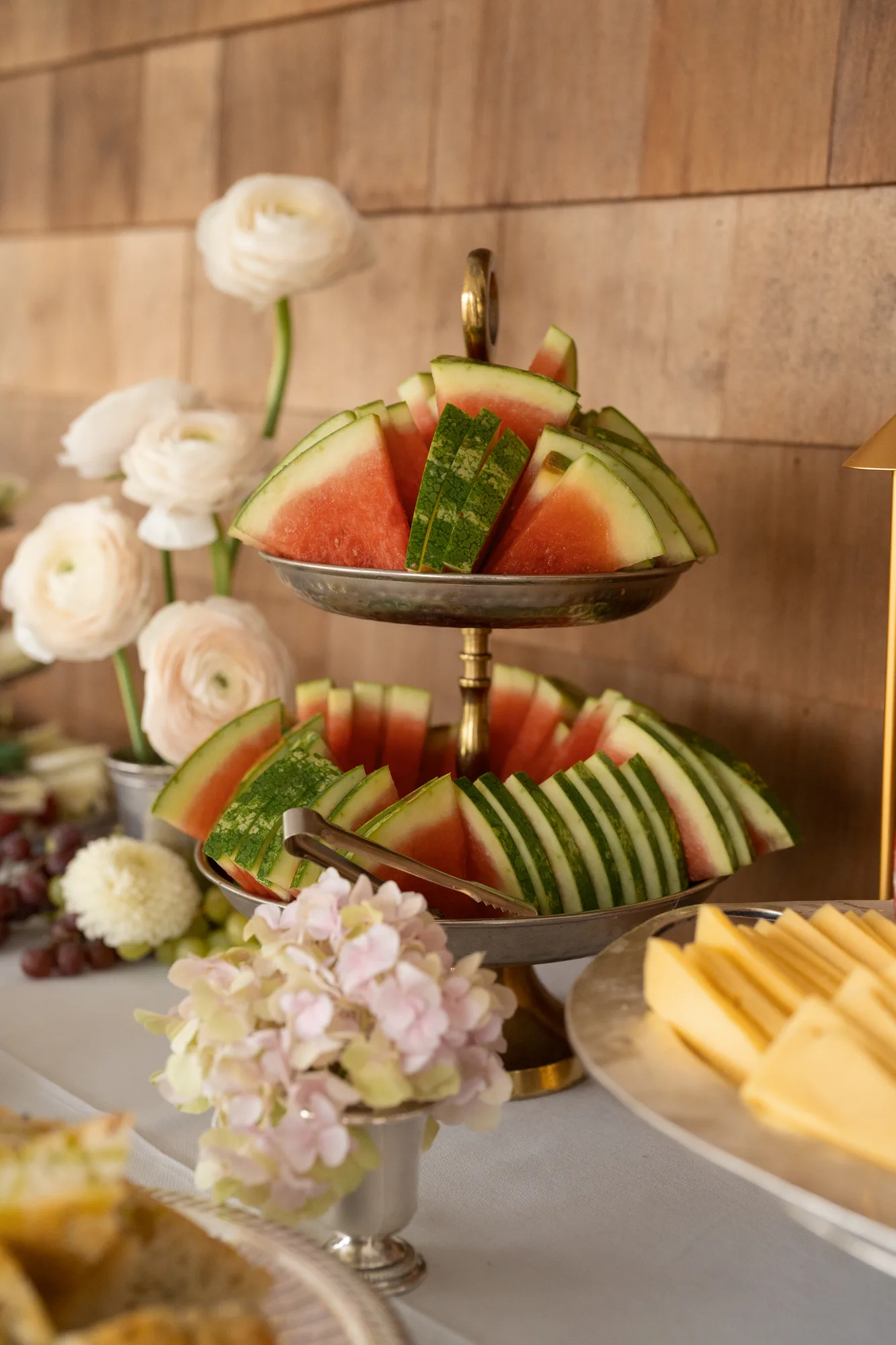 A tiered brass stand of watermelon slices with blush ranunculus behind and a silver cup of faded hydrangea in front