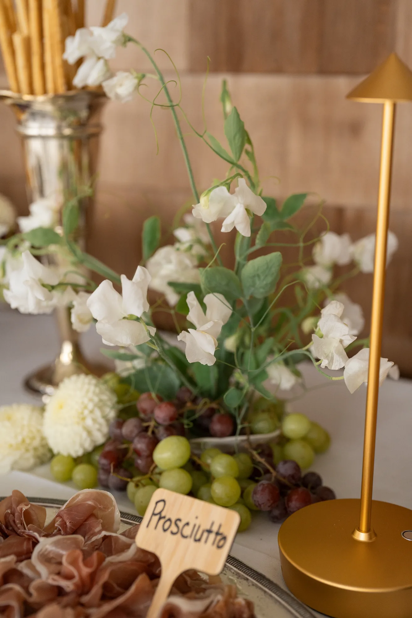 White sweet pea on trailing tendrils in a low dish among grapes on the buffet, a brass lamp beside it