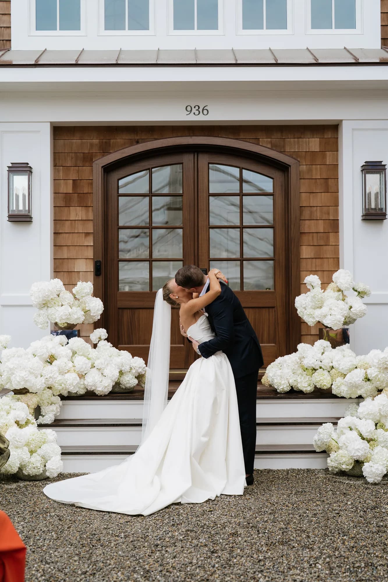 Lindsey and Finn kiss on the gravel before the front steps, white hydrangea banked on both sides