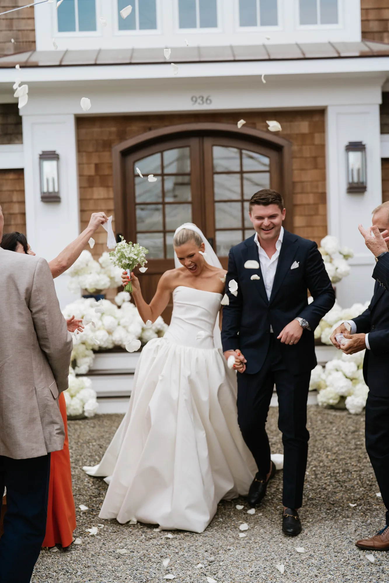 Lindsey raises her tweedia bouquet as guests toss white petals, Finn laughing beside her on the gravel