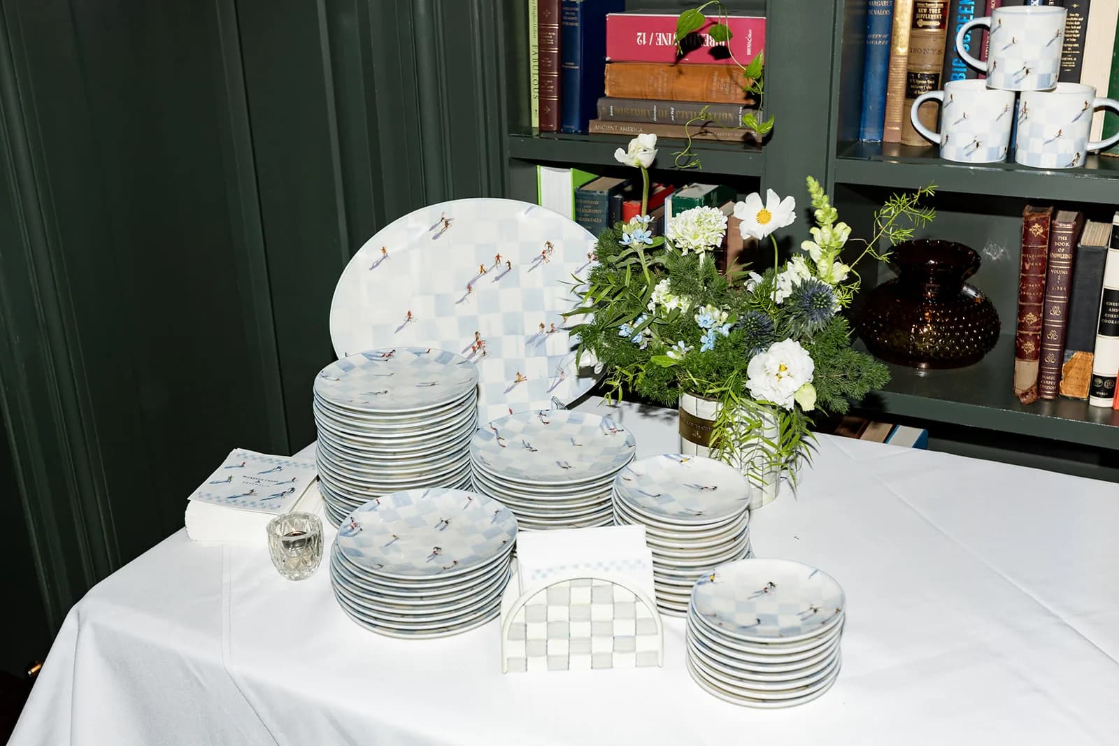 Stacked MacKenzie-Childs ski-print plates beside a white and blue arrangement in a check canister against green shelves