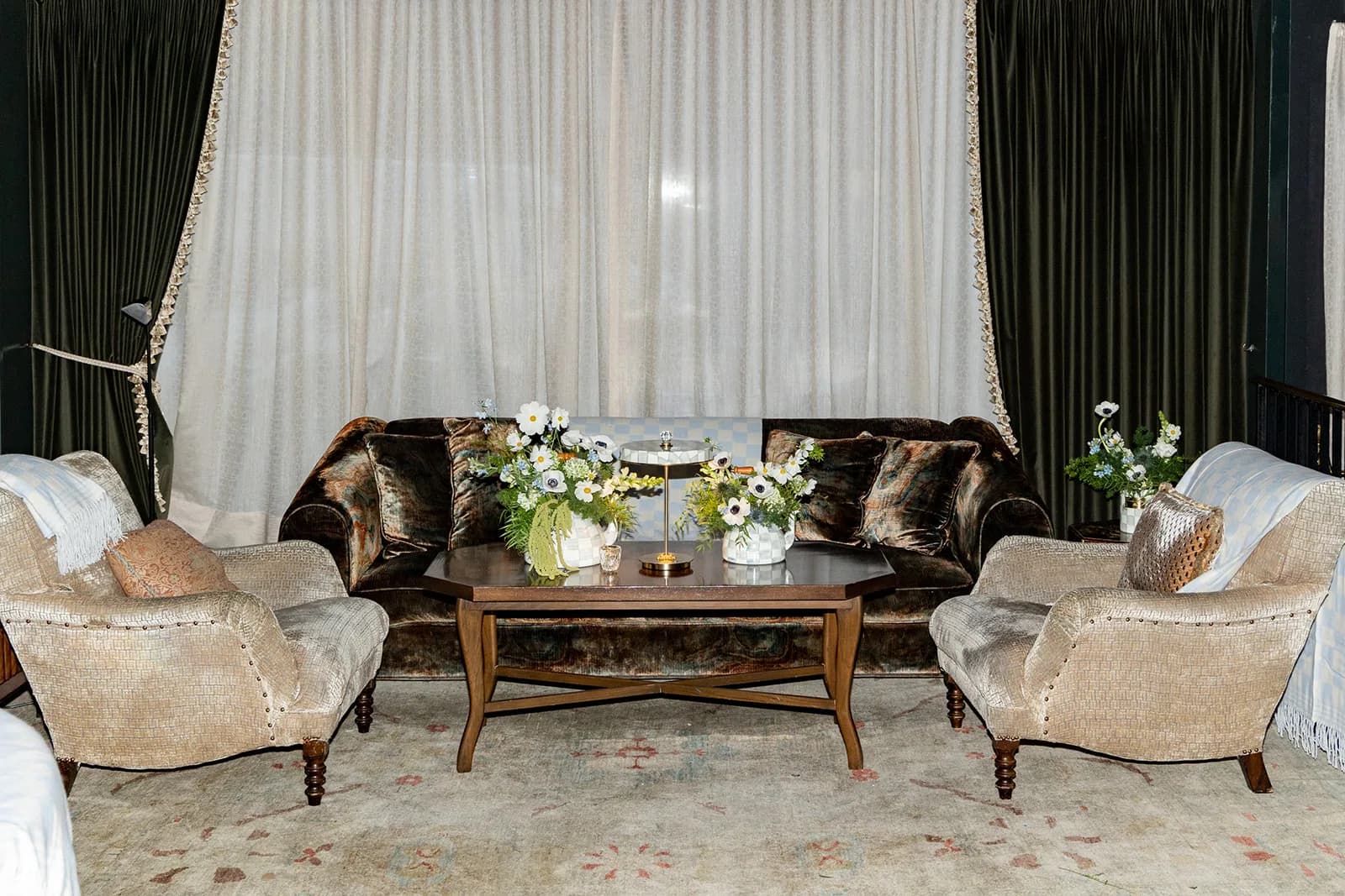 Two arrangements of cosmos and anemones in check pots flank a small lamp on a coffee table between velvet sofa and armchairs