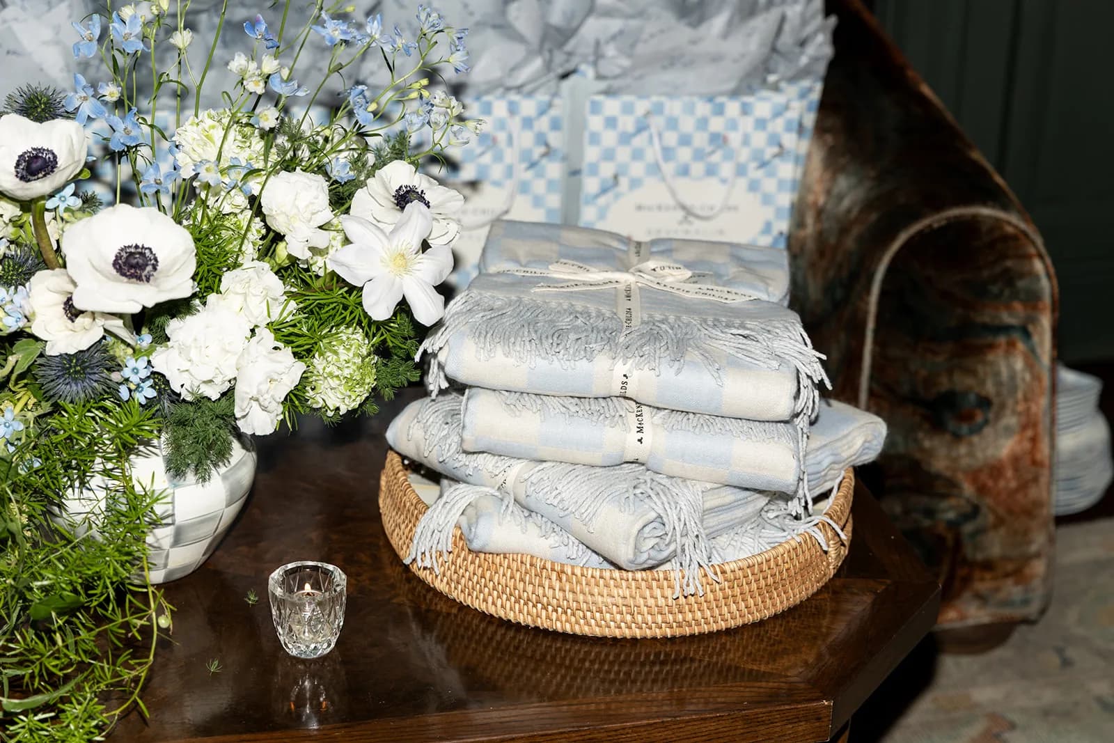 Anemones and blue delphinium in a check canister beside folded MacKenzie-Childs throws stacked in a rattan tray