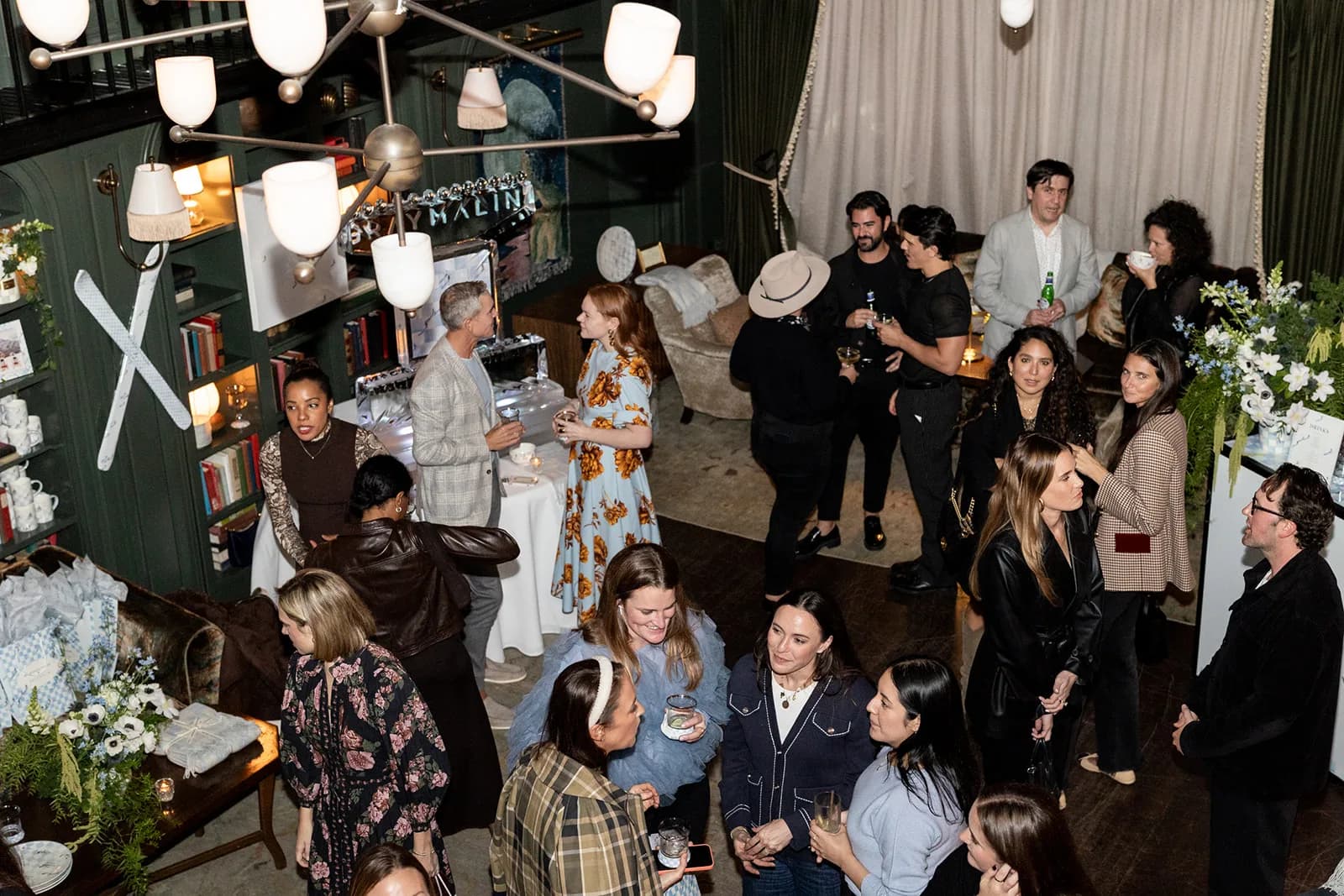 Guests mingle beneath a brass chandelier in the green library lounge, arrangements and gift bags at the edges of the room
