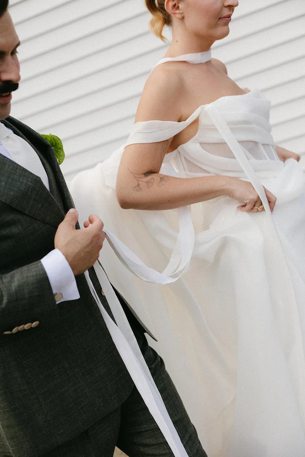 Bride and groom walk along white clapboard, a green anthurium boutonniere on his lapel and her sash ribbons in his hand