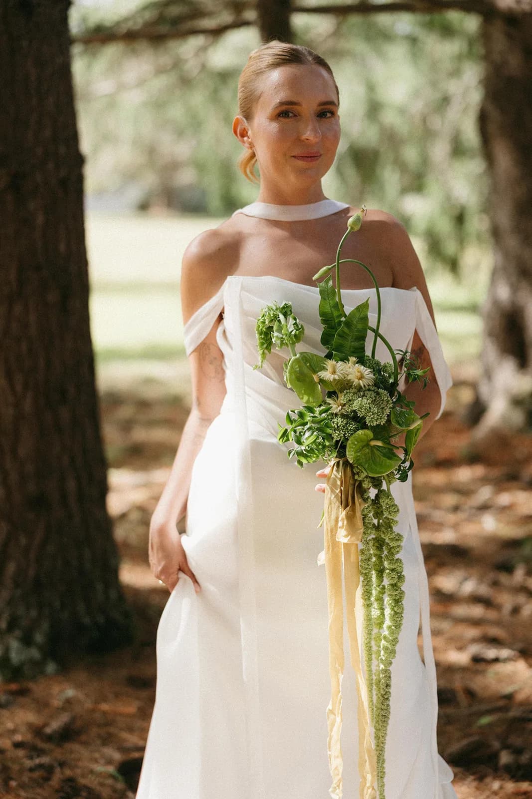 Bride stands among pines holding a sculptural green bouquet, two allium stems curling above trailing amaranthus