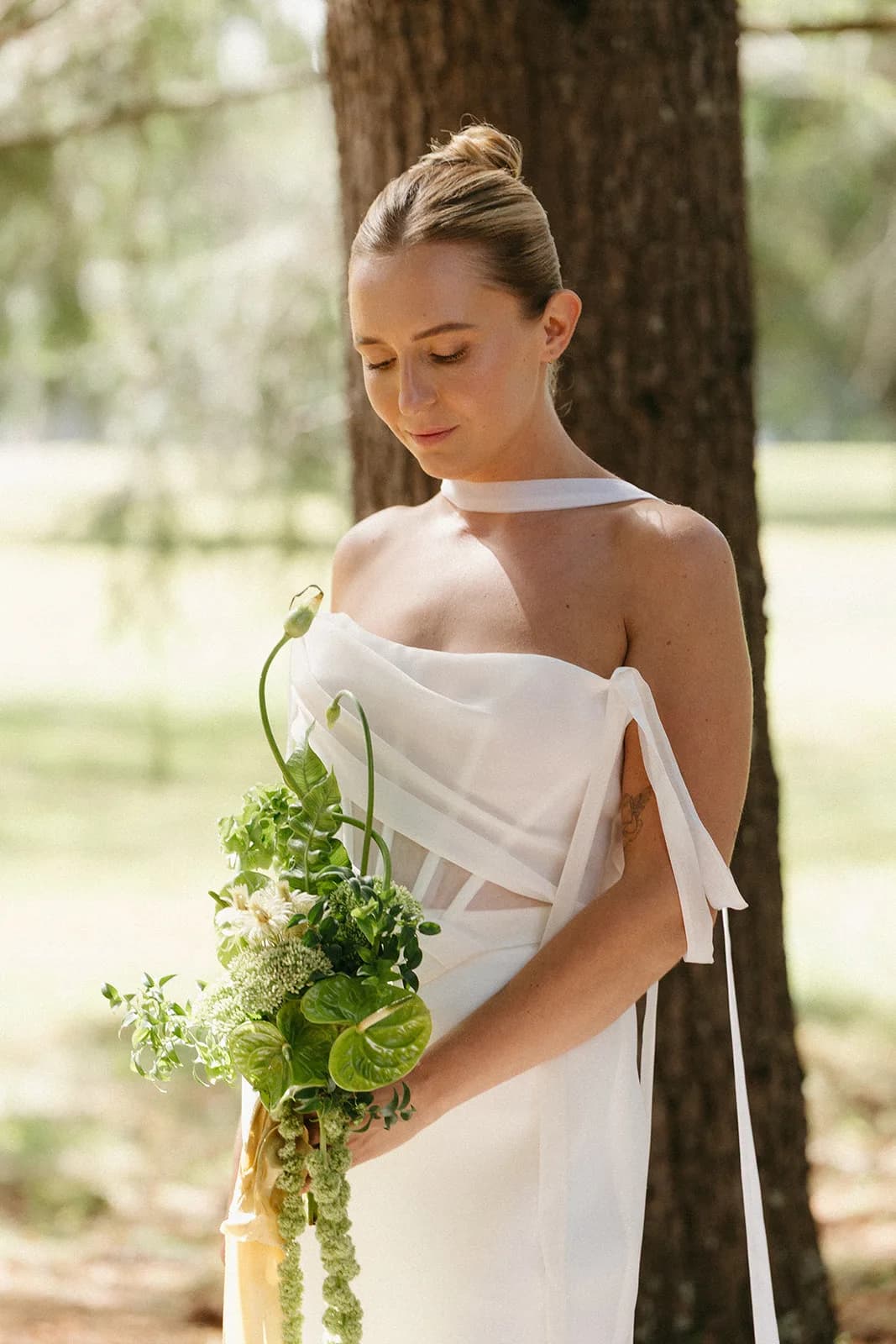 Bride looks down at her bouquet of anthurium, bells of Ireland, and curling allium stems in dappled light beneath a pine
