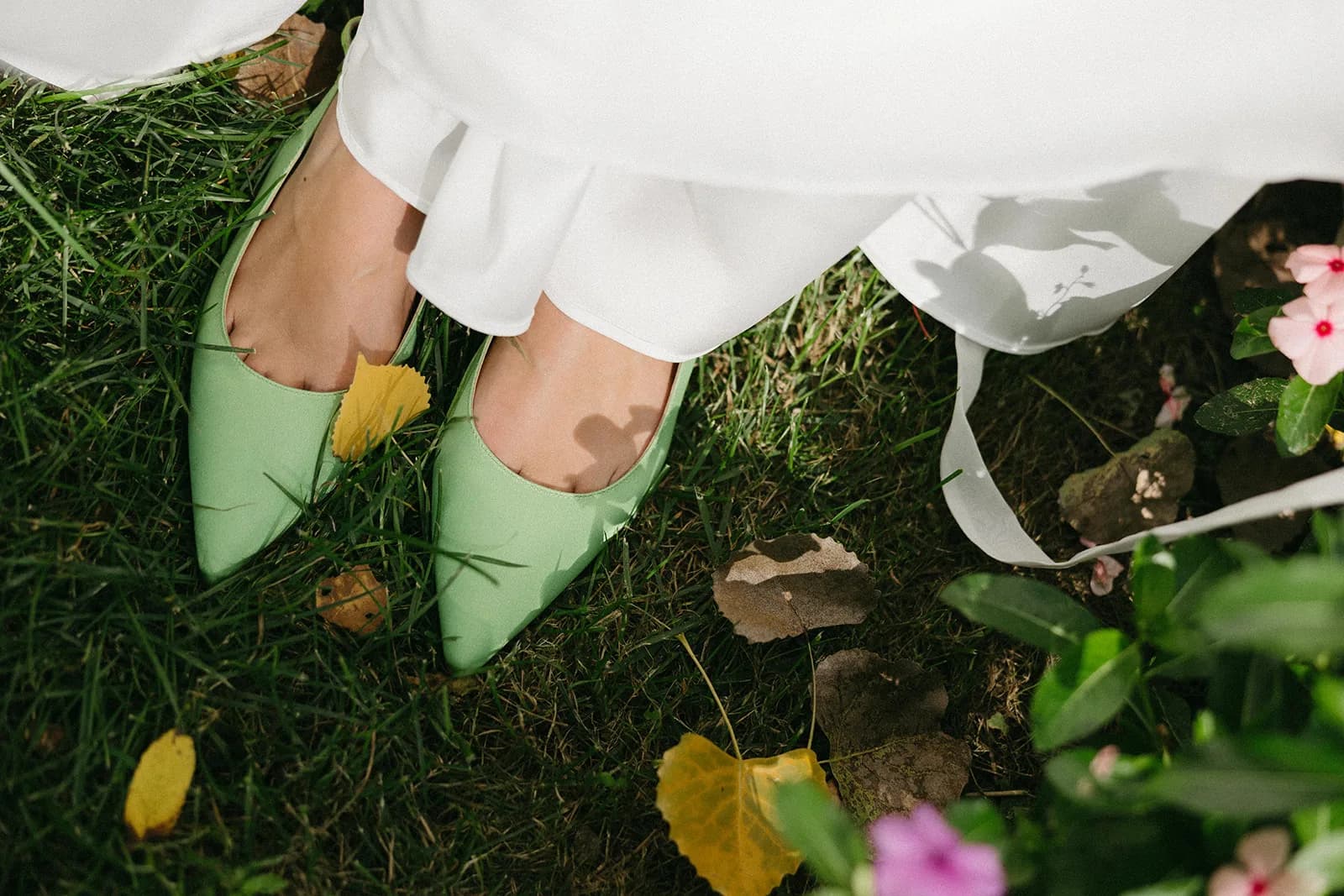 Bride's sage green pointed flats on the lawn beside her dress hem, fallen yellow leaves, and a trailing white ribbon
