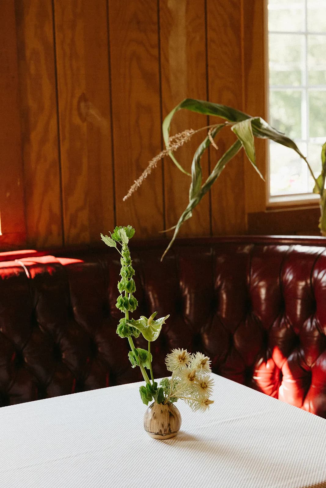 Ikebana-inspired bud vase on a white tablecloth, bells of Ireland rising past a single anthurium by a red leather banquette