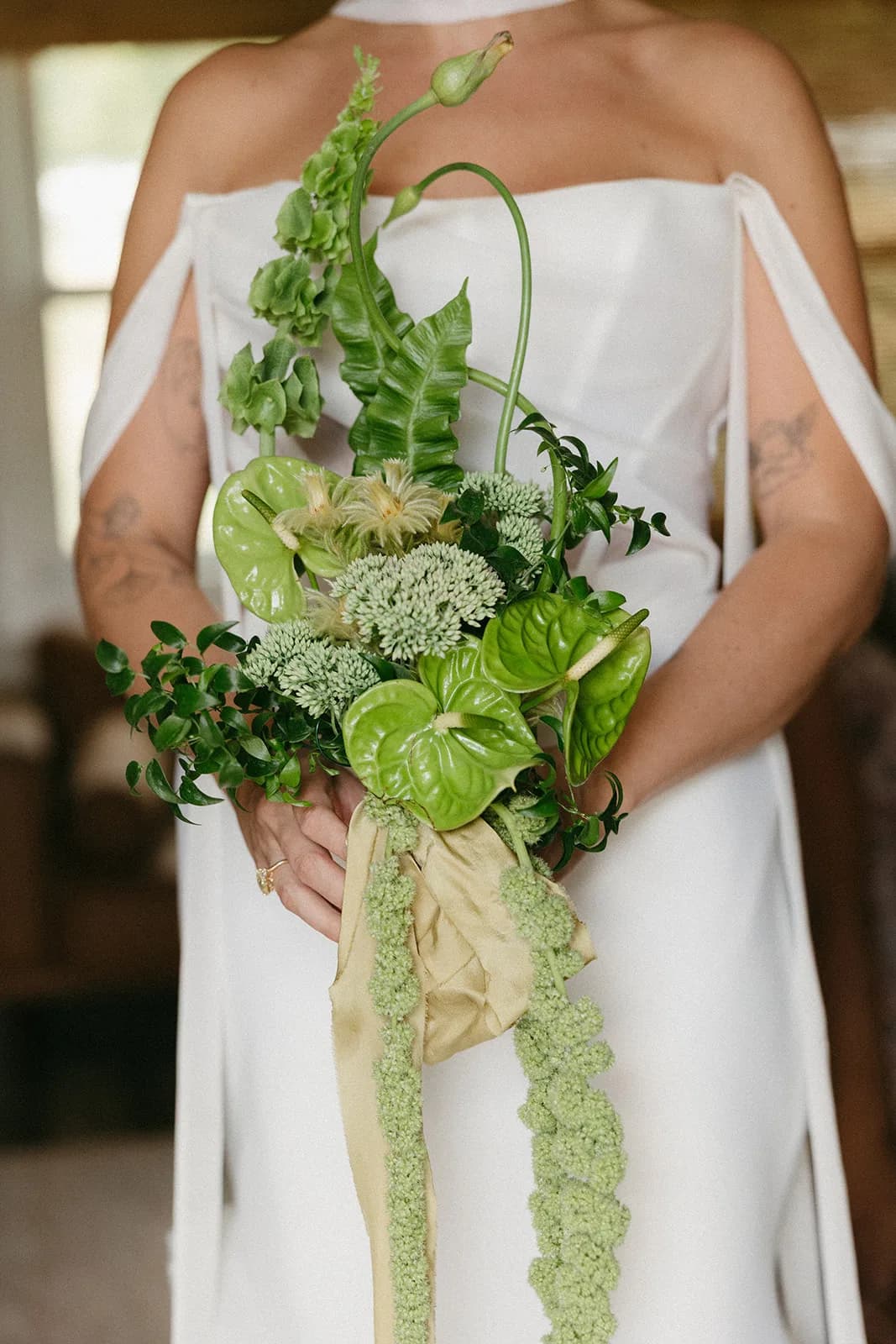 Bride holds a bouquet of green anthurium, bells of Ireland, Phylica, and two curling allium stems with gold silk ribbon