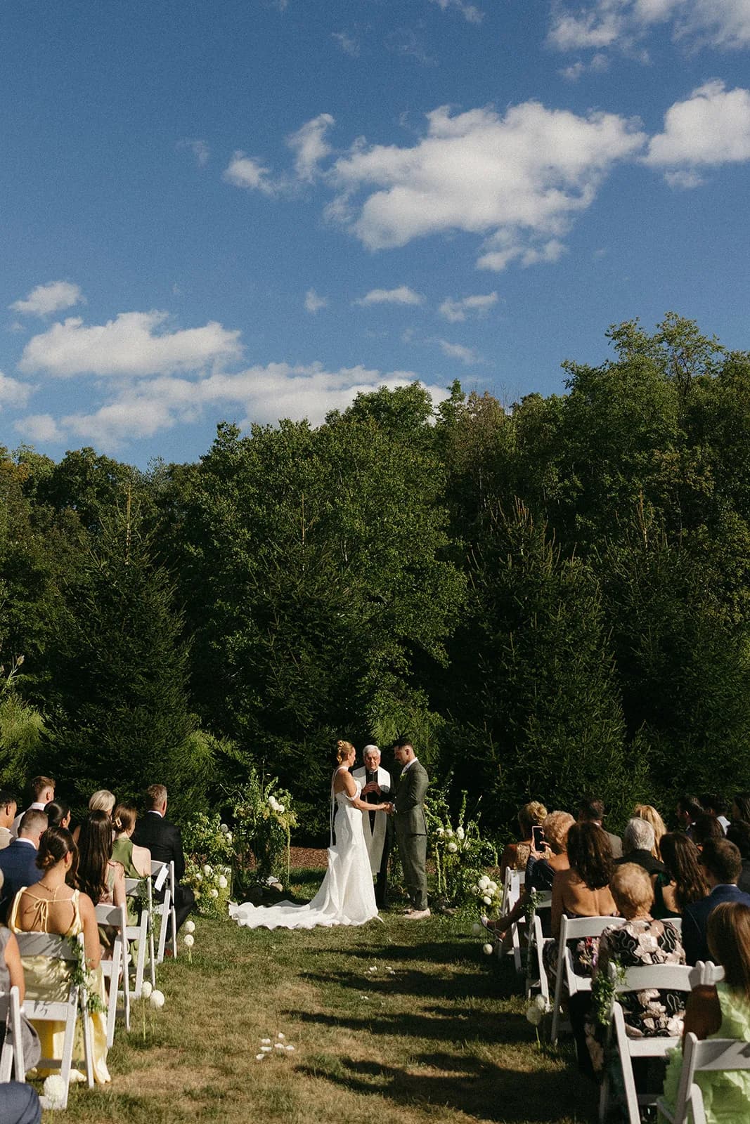 Wide view of the ceremony under blue sky, couple before pines flanked by low green arrangements and single dahlias on chairs