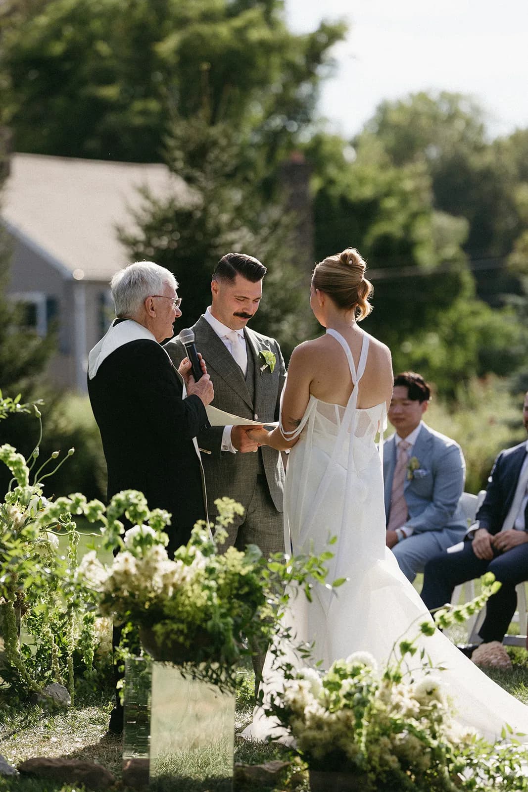 Couple hold hands as the officiant reads, ground arrangements of bells of Ireland, white dahlias, and trailing amaranthus