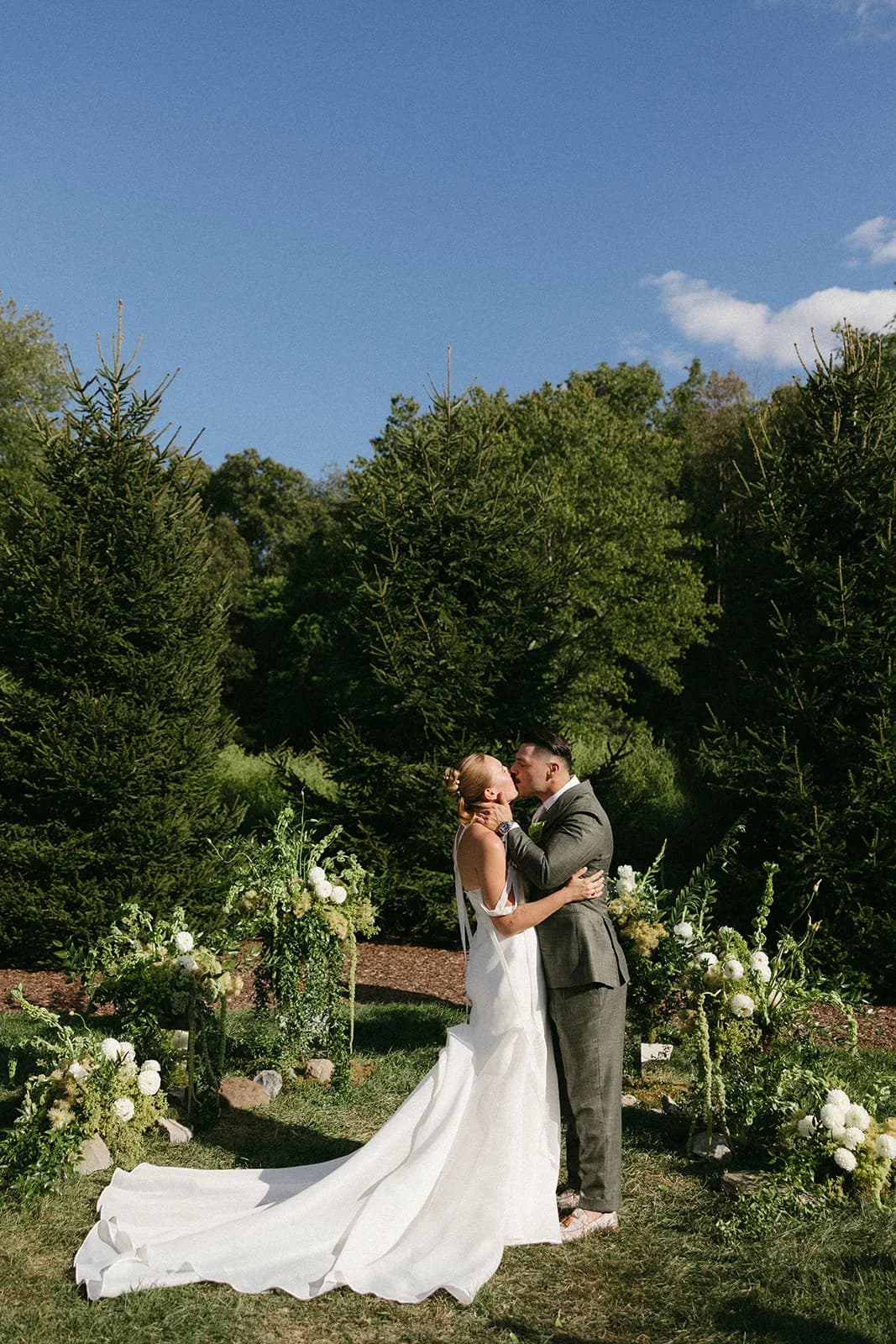 First kiss before a wall of pines, low sculptural arrangements of white dahlias and trailing amaranthus framing the couple