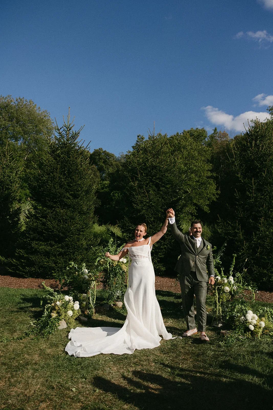 Couple raise joined hands in the recessional, white dahlia arrangements with trailing amaranthus on the grass behind them